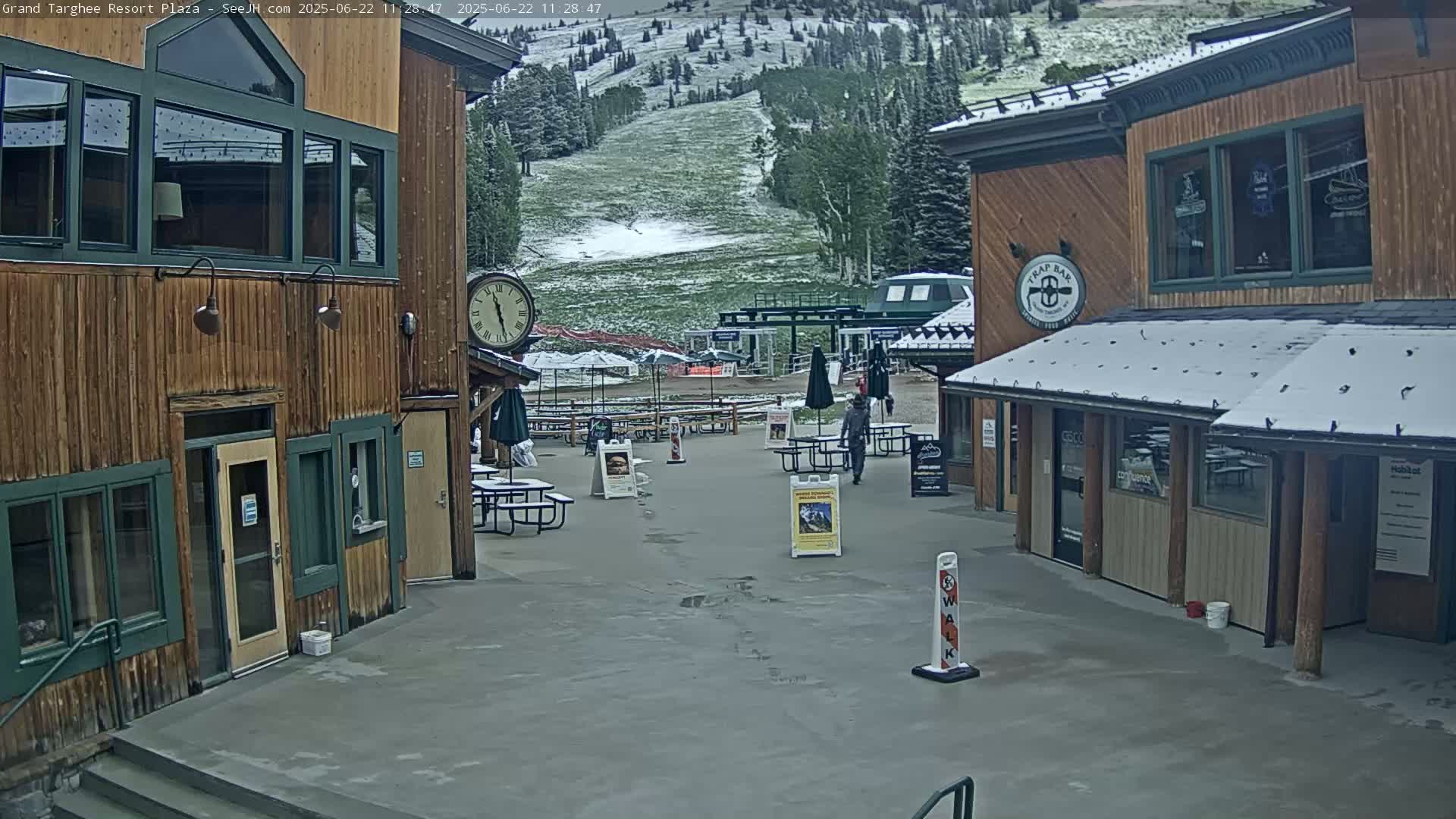 A plaza with wooden buildings, partially covered in snow, overlooks a snow-dusted ski slope under a cloudy sky.