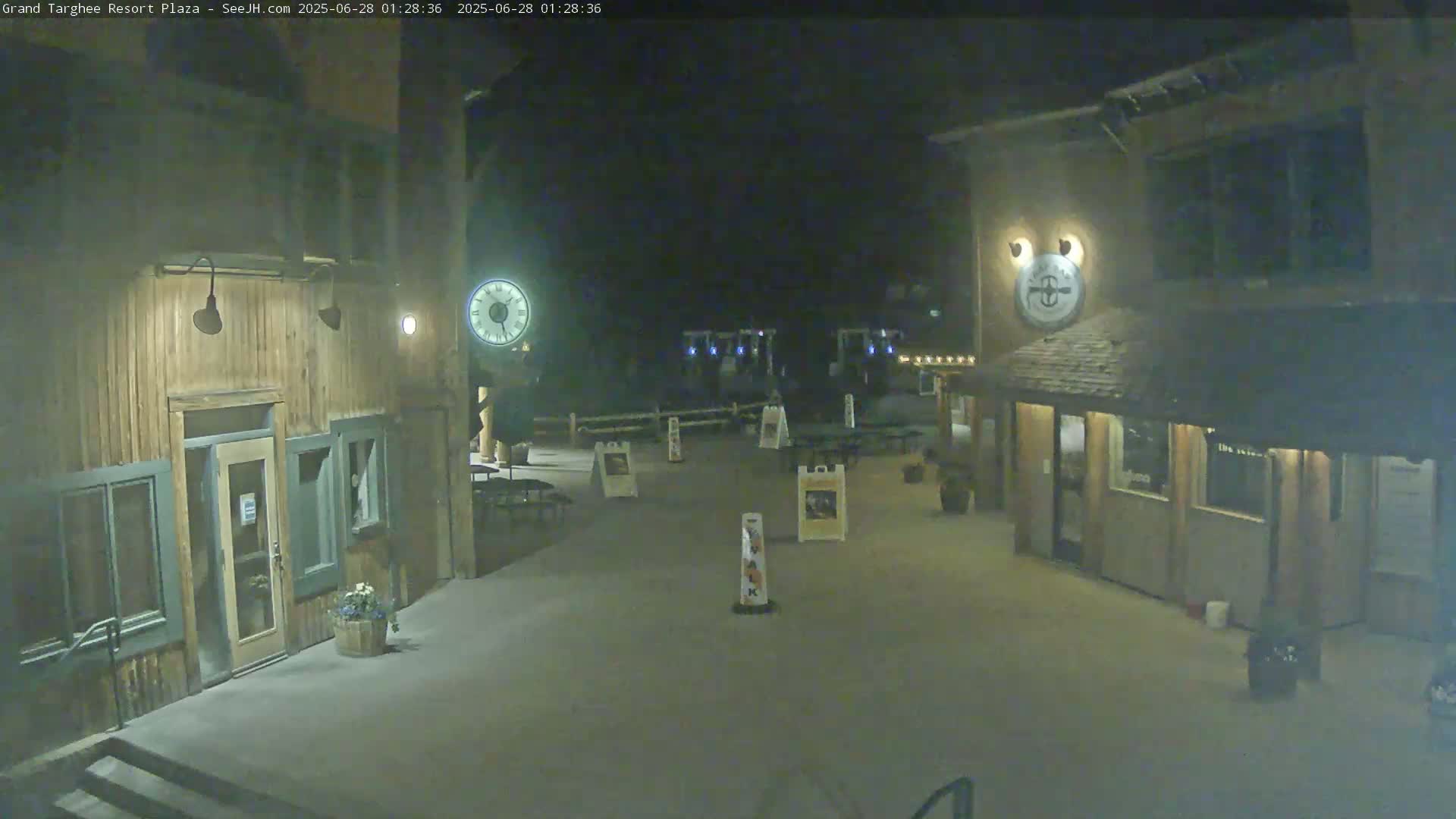 A nighttime view of a plaza with two wooden buildings, outdoor seating, and a clock.