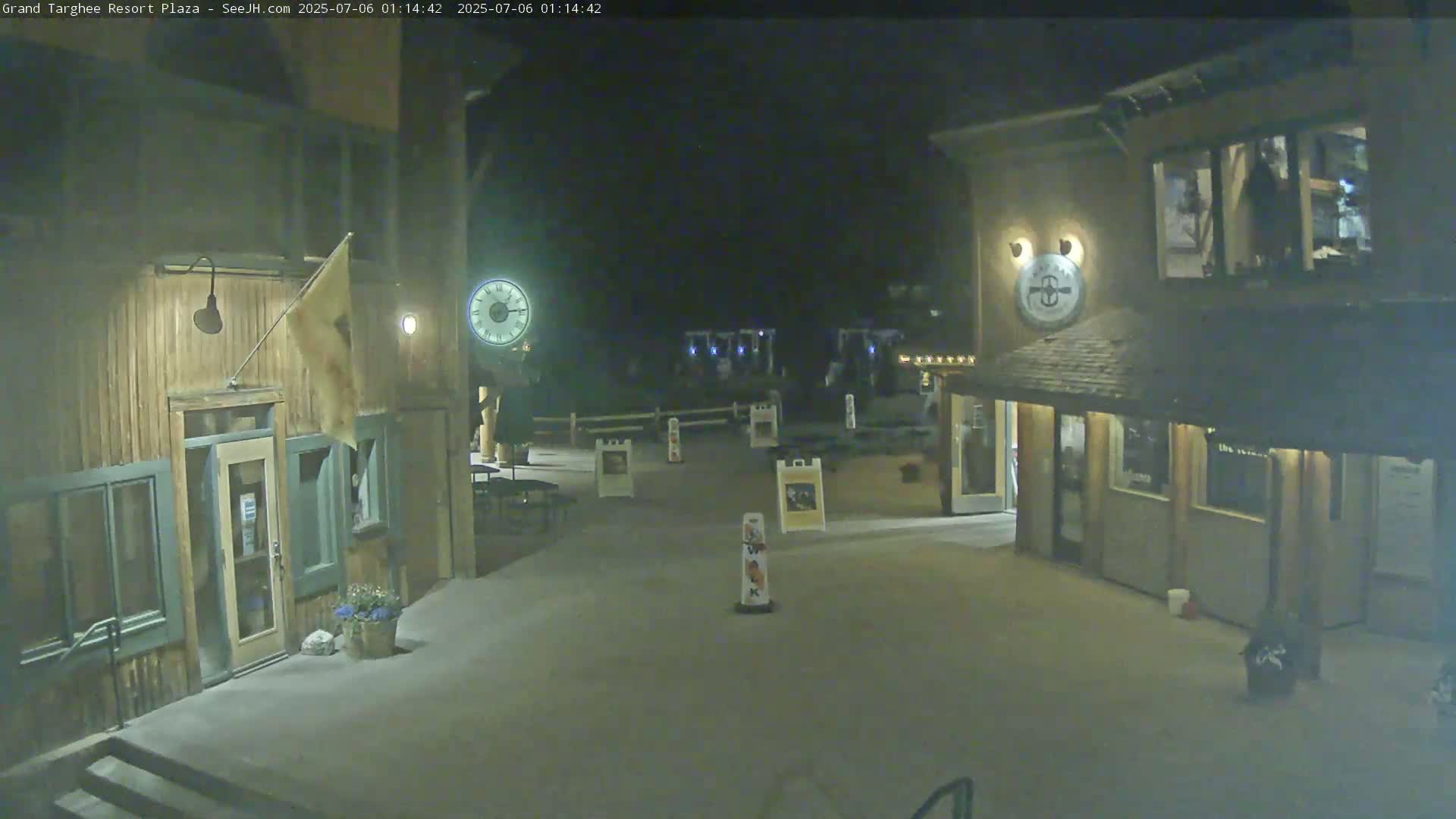 A nighttime view of a plaza with two wooden buildings, a clock, and several signs.