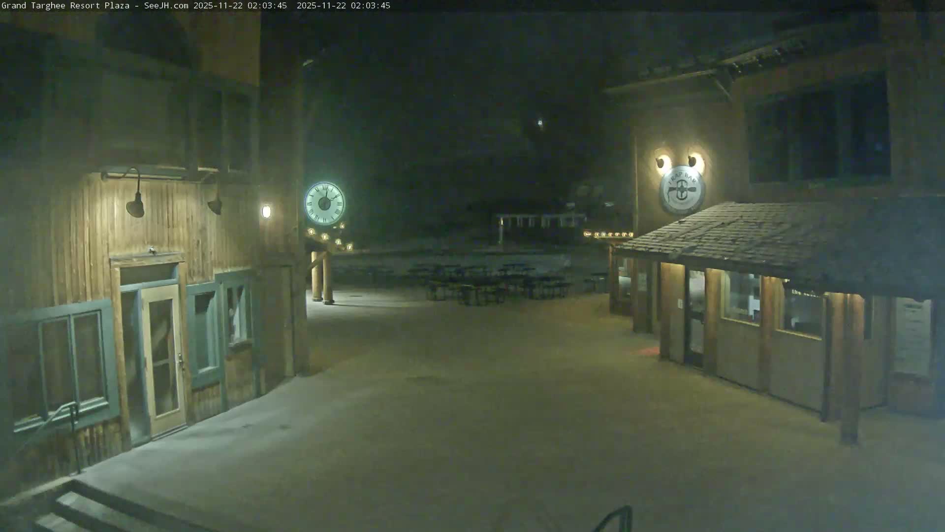 The image depicts a quiet, dimly lit outdoor resort plaza at night, featuring rustic wooden buildings, a prominent clock, and numerous empty picnic tables on a clear, dry paved surface under a dark sky.