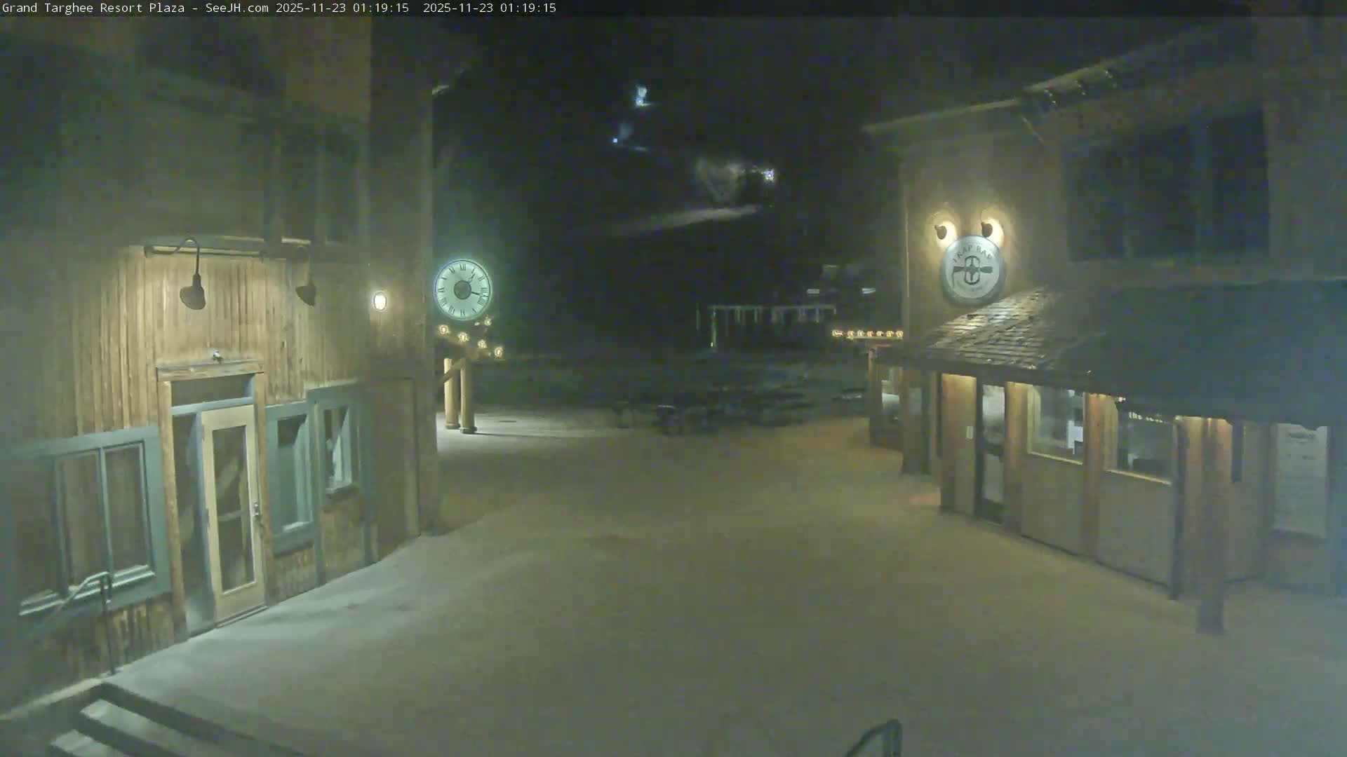 A quiet outdoor plaza at a resort is illuminated by warm lights from rustic wooden buildings and streetlights on a clear night with a thin layer of snow covering the ground.
