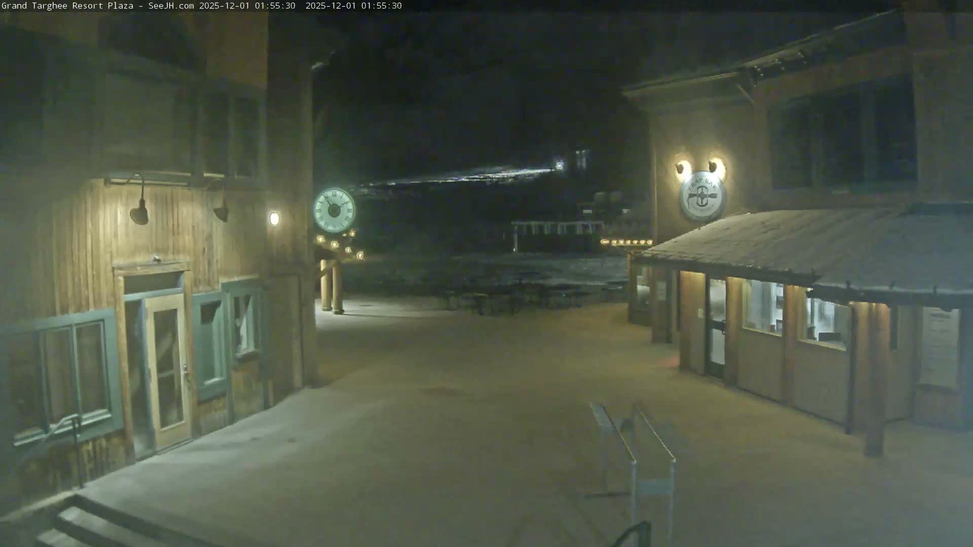 A quiet, snow-dusted resort plaza is illuminated at night by warm lights from rustic buildings and an outdoor clock, with distant slope lights visible under clear skies.