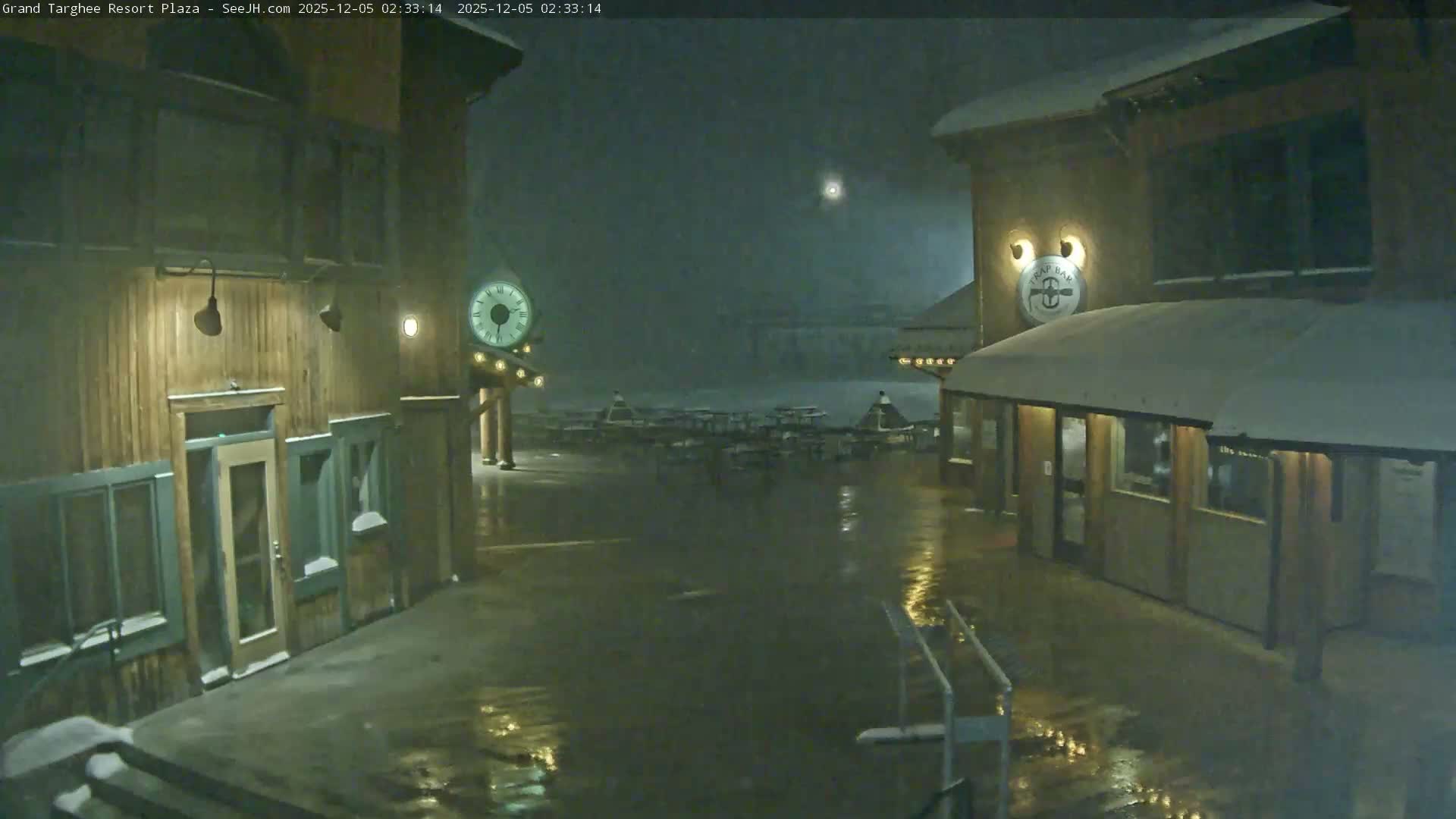 A snow-covered outdoor plaza is seen at night, with warm lights illuminating the wet ground and buildings on either side as snow falls.