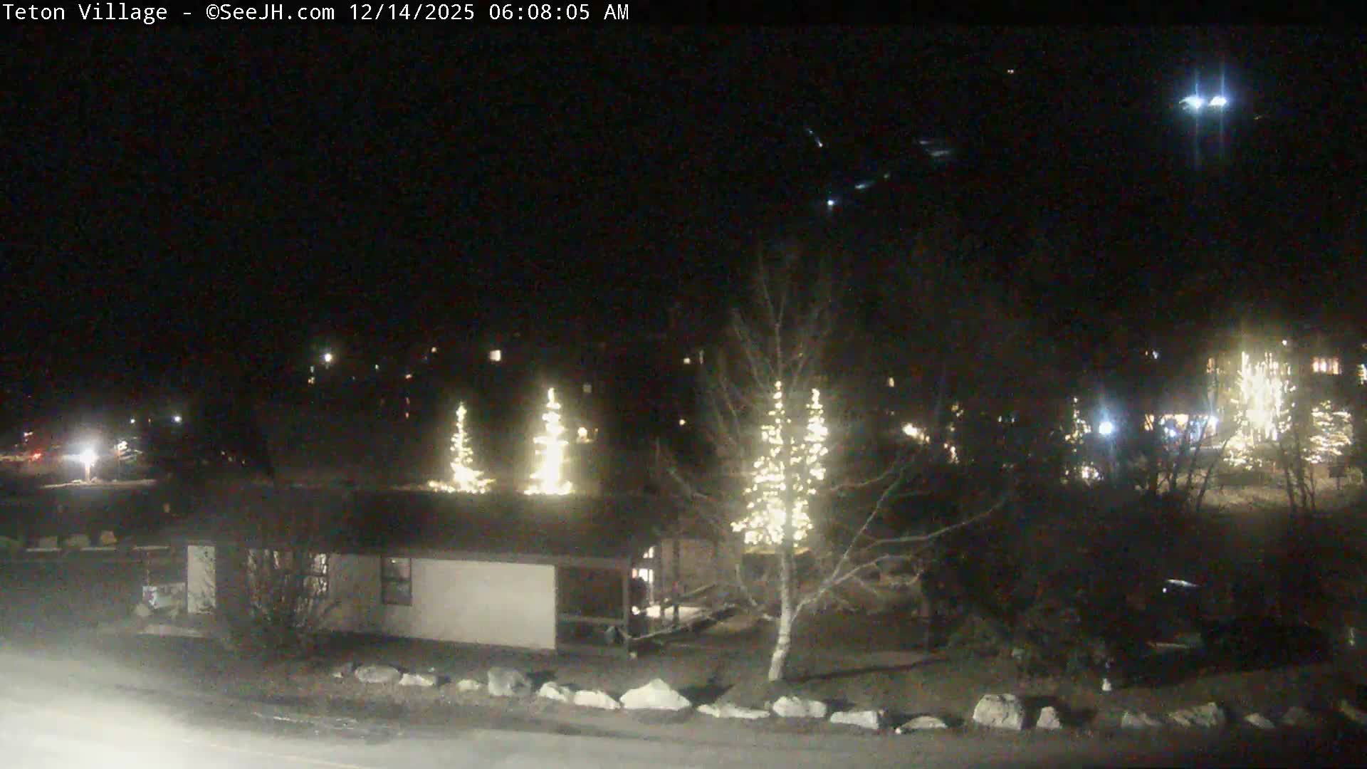 A snow-covered outdoor plaza is seen at night, with warm lights illuminating the wet ground and buildings on either side as snow falls.