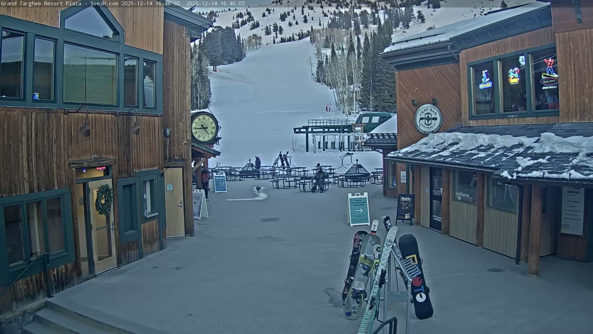 A snow-covered outdoor plaza is seen at night, with warm lights illuminating the wet ground and buildings on either side as snow falls.