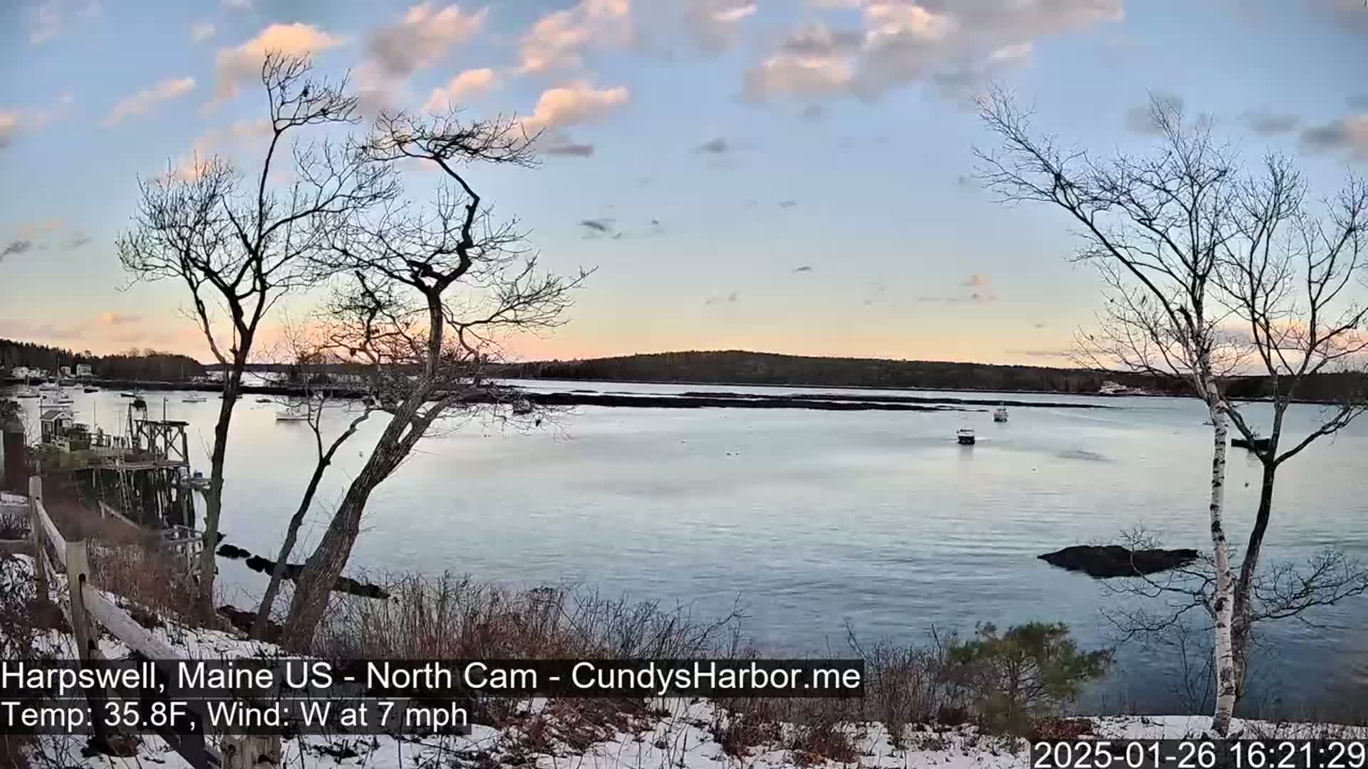 Here is a description of the image: A tranquil waterfront scene at sunset, with a calm body of water dotted with a few small boats, under a partly cloudy sky, and a light dusting of snow on the ground in the foreground.