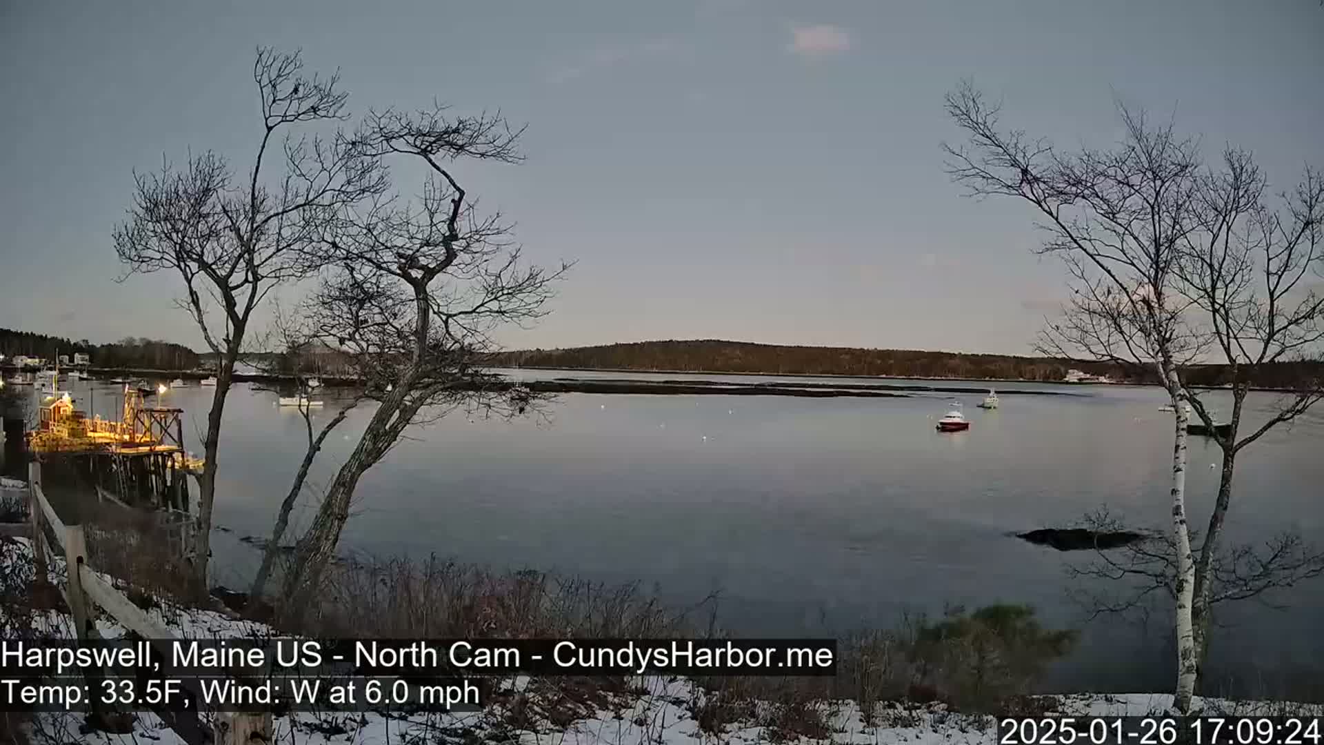Here's a description of the image: A calm body of water, with a few boats visible, is seen from a snow-dusted shoreline under a twilight sky, with bare trees in the foreground.