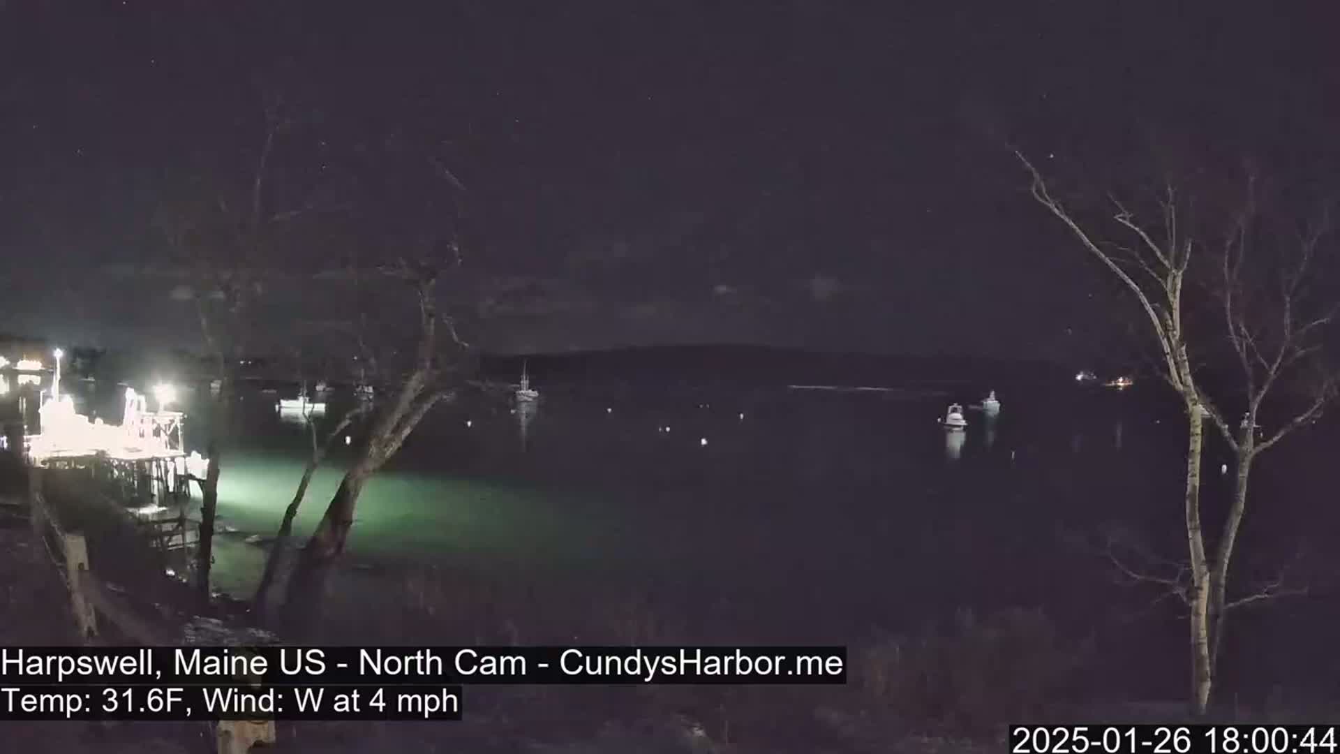 Here is a description of the image: A nighttime view of a calm body of water with several boats visible, under a dark, clear sky with a few stars, and a temperature of 31.6°F and a west wind at 4 mph.