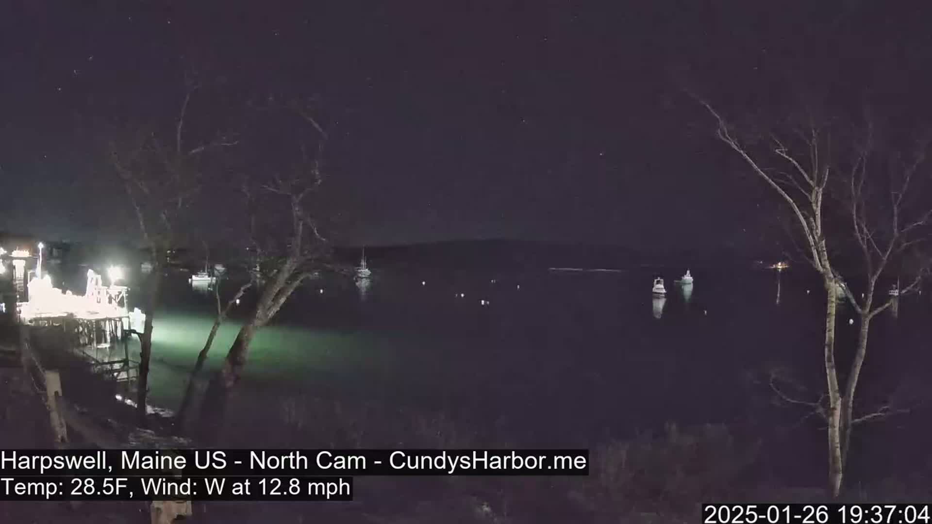 A nighttime view of a calm body of water with several boats moored, under a clear, starry sky with a westerly wind of 12.8 mph and a temperature of 28.5°F.