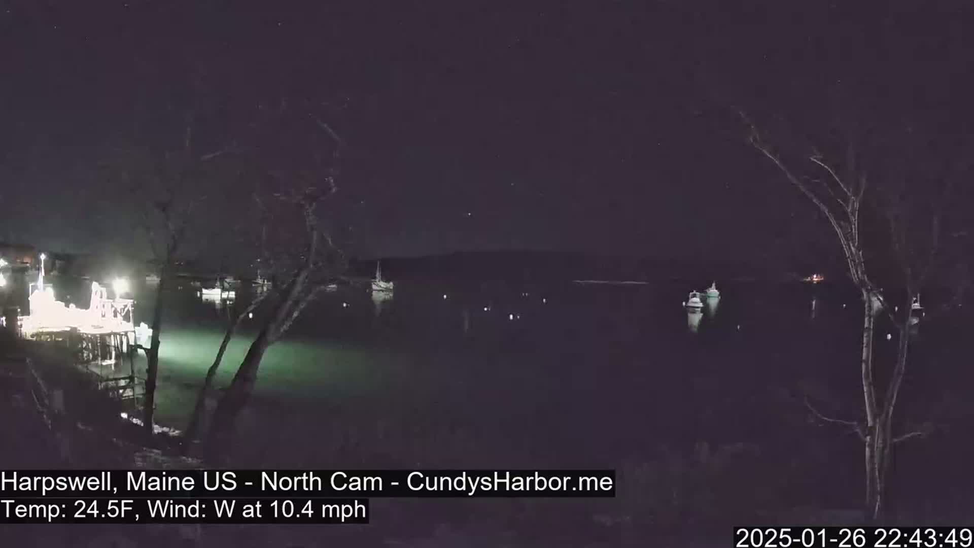 At night, under a clear sky with a west wind of 10.4 mph and a temperature of 24.5°F, several boats are moored in a calm harbor, illuminated by lights on a nearby dock.