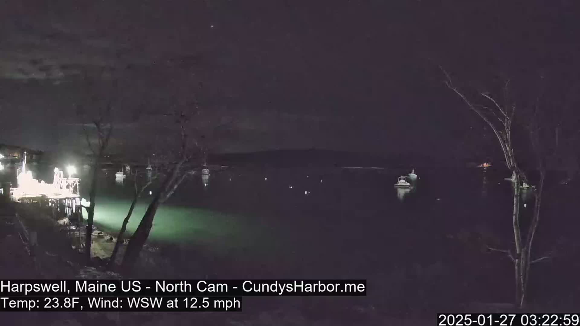 A nighttime view of a harbor with several boats moored, under a clear, dark sky with a 23.8°F temperature and a 12.5 mph wind from the west-southwest.