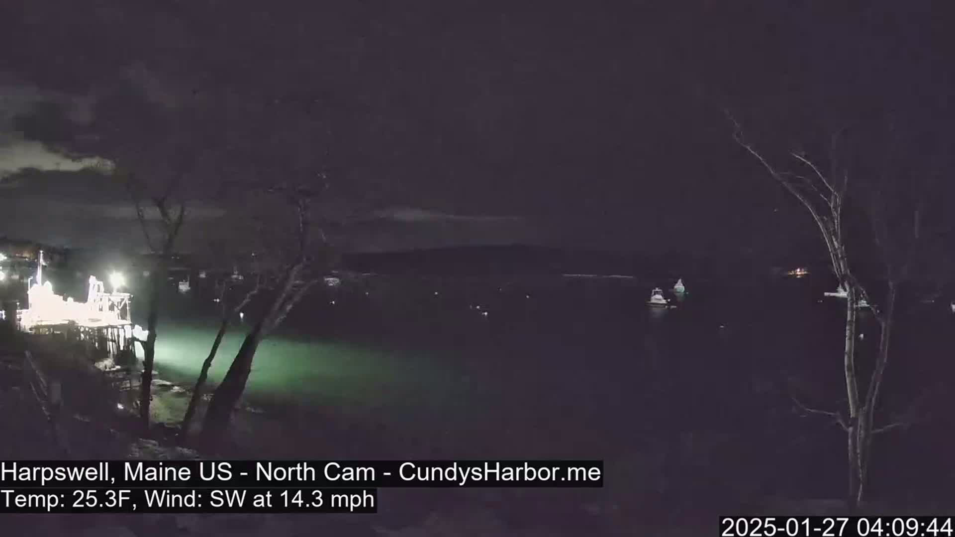 A nighttime view of a harbor with several boats, under a dark sky with a southwest wind at 14.3 mph and a temperature of 25.3°F.