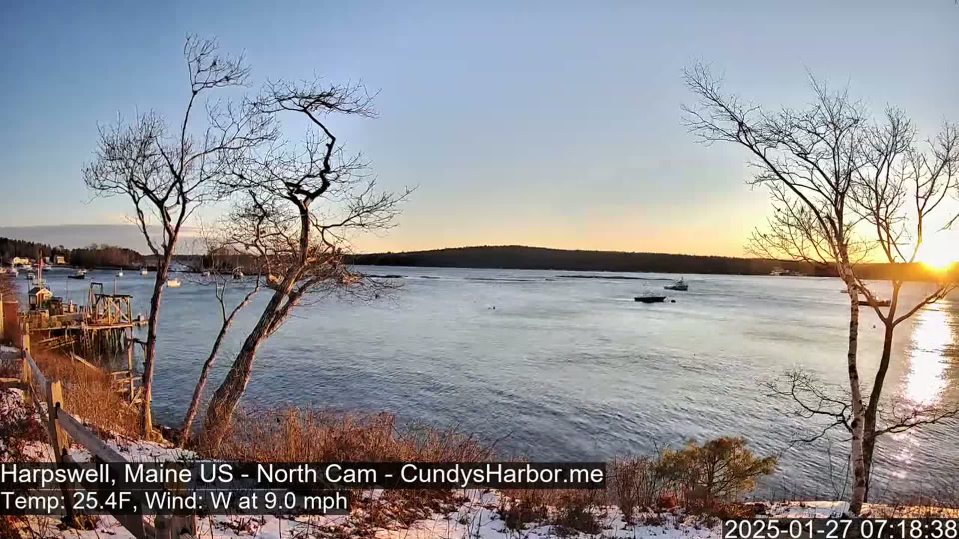 Here is a description of the image: A tranquil waterfront scene at sunset, with bare trees in the foreground and a calm body of water dotted with a few boats, under a clear sky with the sun setting on the horizon; there is a light dusting of snow on the ground.