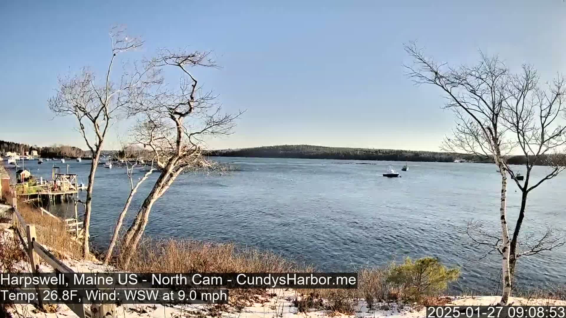 A calm, sunny day on a body of water, with a few boats and leafless trees in the foreground, and a snow-dusted shoreline.