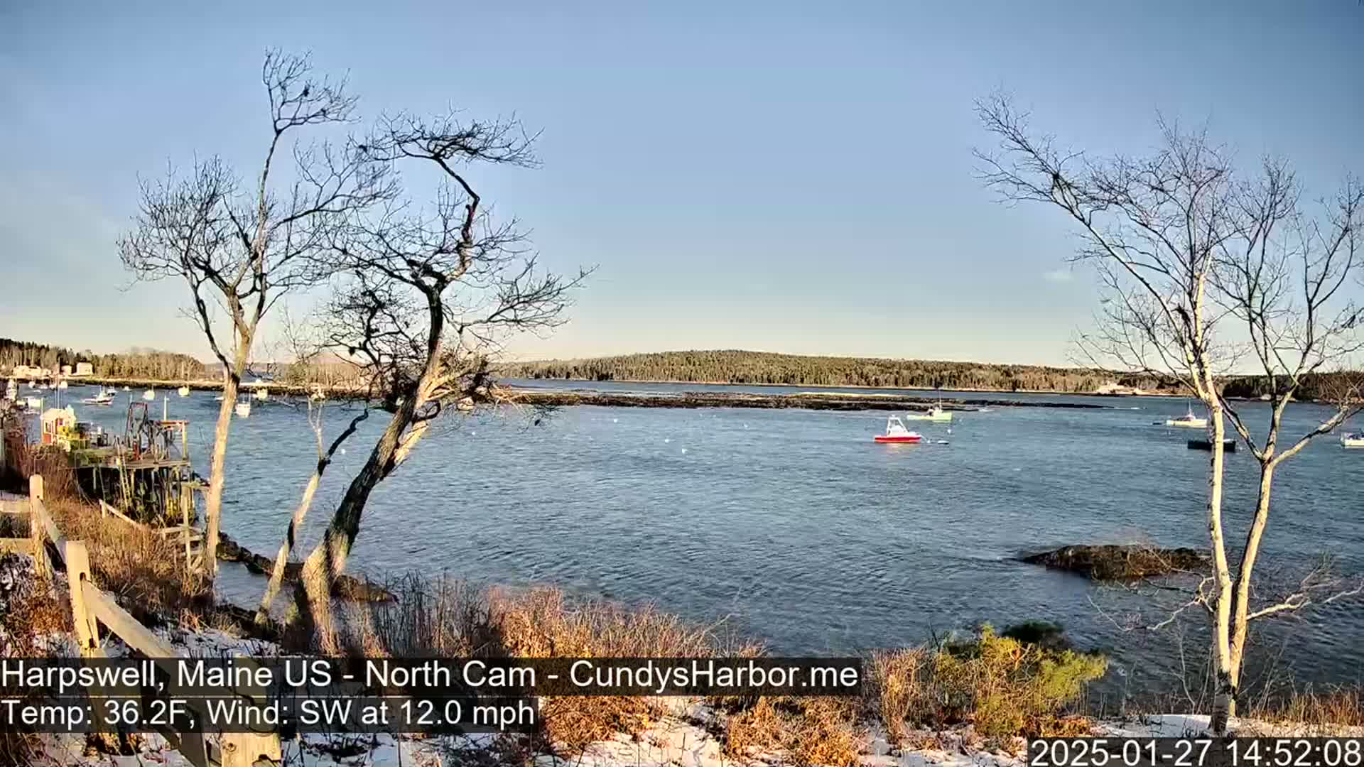 A calm body of water with several boats is viewed from a snow-dusted shoreline under a clear, sunny sky.