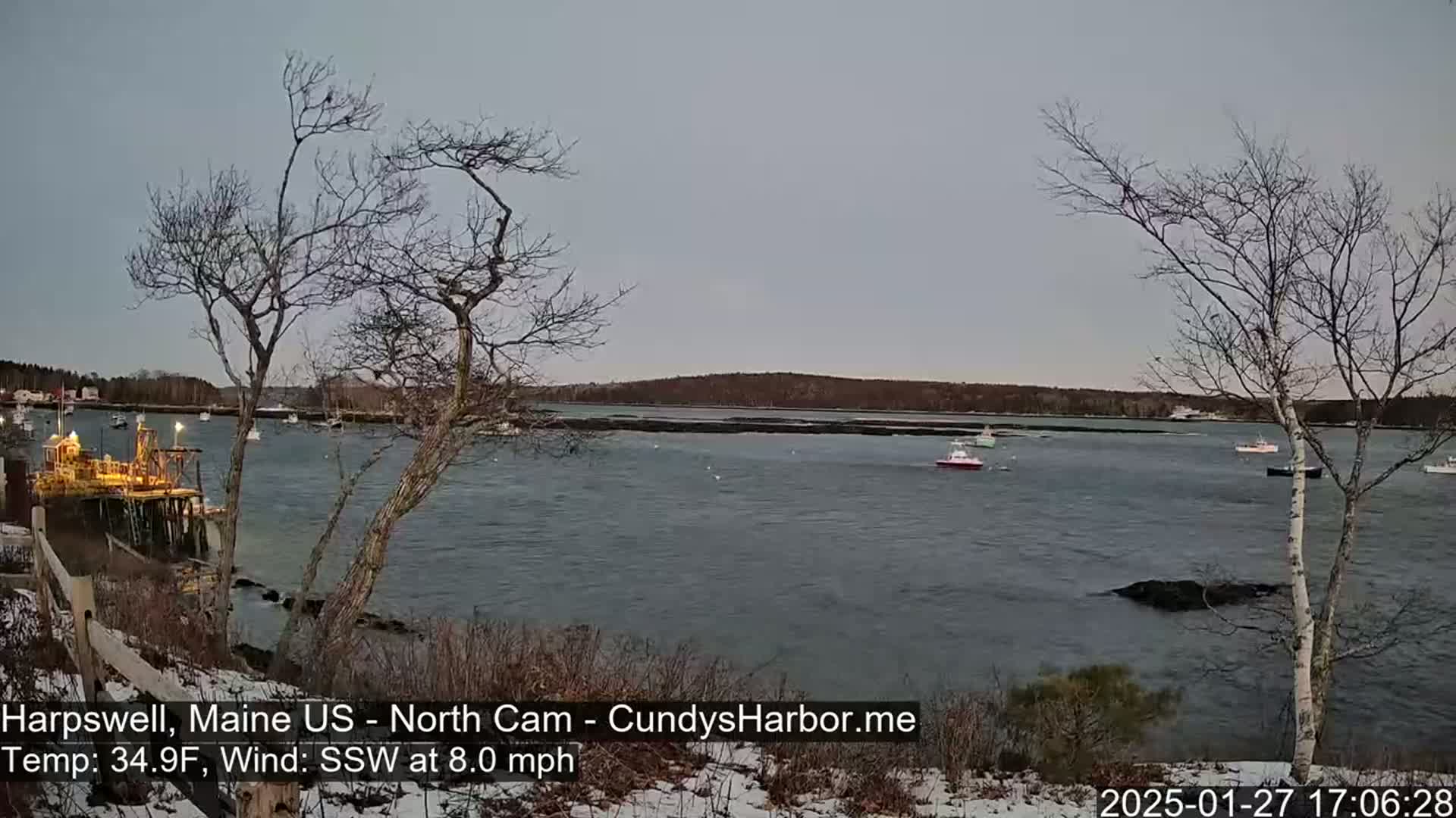 A calm body of water with several small boats is viewed from a snow-dusted shoreline under a gray sky, with bare trees in the foreground.