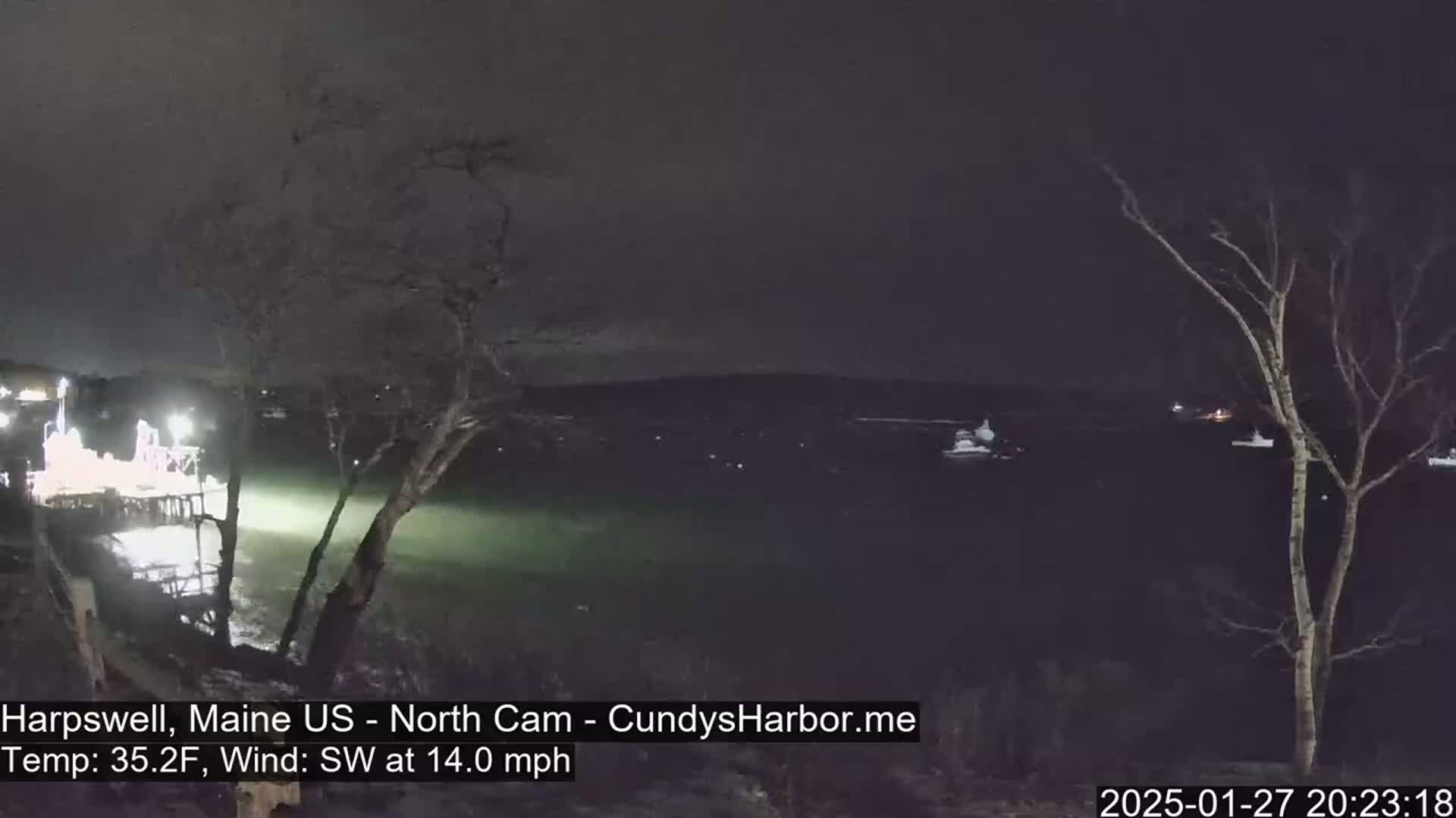 A nighttime view of a dark body of water with a few boats visible, a lit pier in the foreground, and bare trees under a dark sky with a southwest wind blowing at 14 mph and a temperature of 35.2°F.
