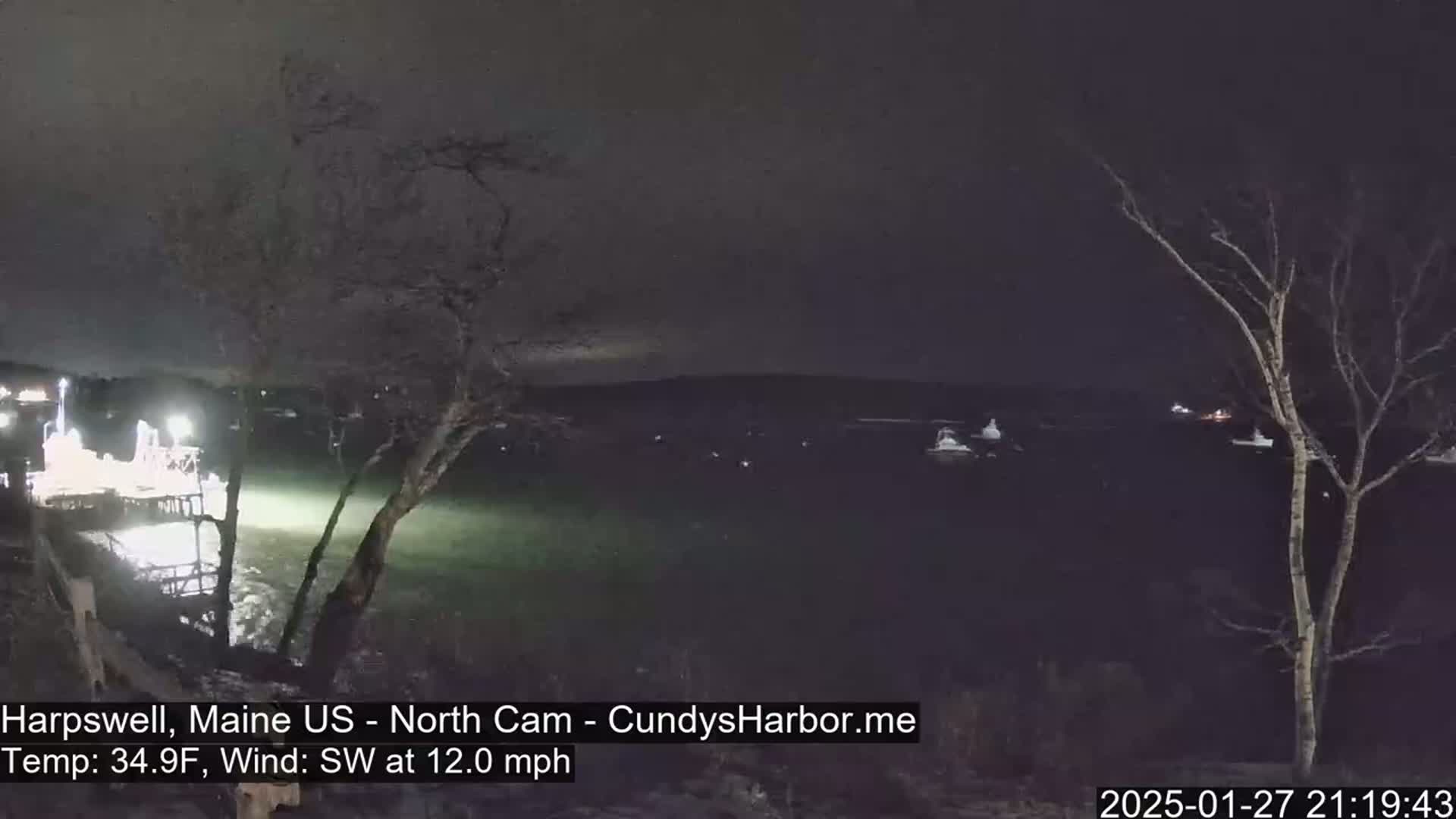 A nighttime view of a dark body of water with some lights visible on boats and a dock, under a cloudy sky with a southwest wind.