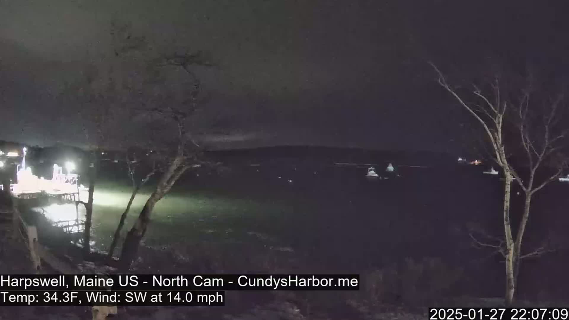 A nighttime view of a dark body of water with a few lights visible on the far shore, under a cloudy sky, with a 14 mph southwest wind.