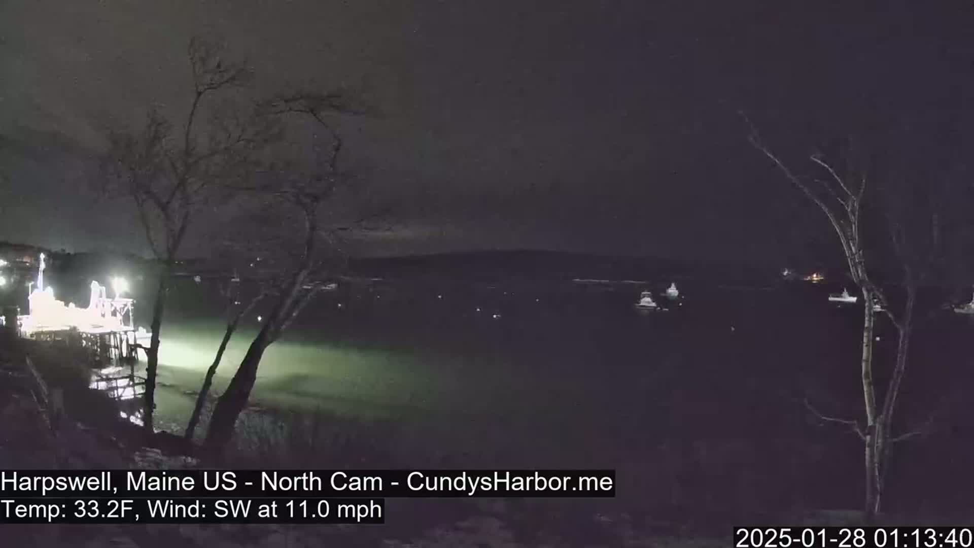 A nighttime view of a calm body of water with several boats and a pier, under a dark sky with a southwest wind at 11 mph and a temperature of 33.2°F.