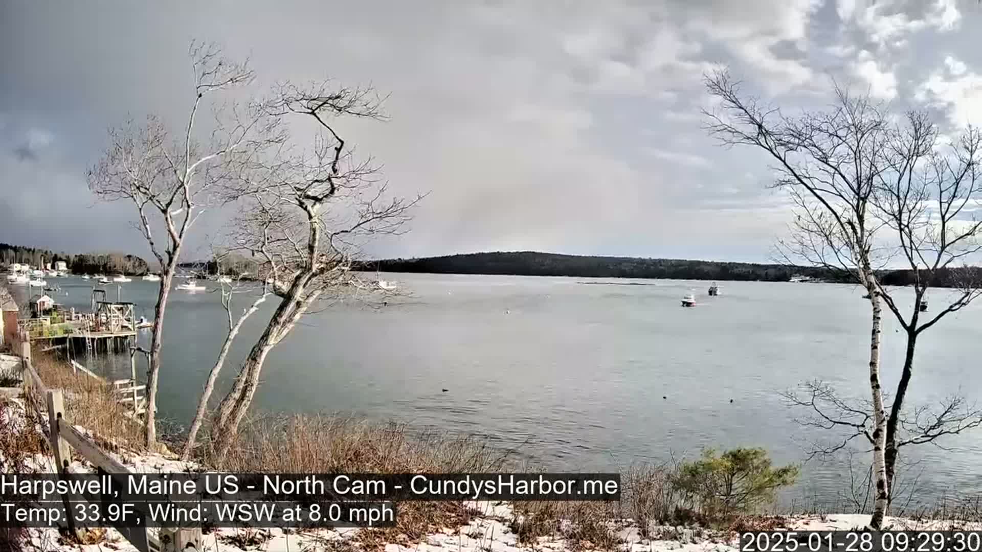 A calm body of water with a few boats is seen from a snow-dusted shoreline under an overcast sky.