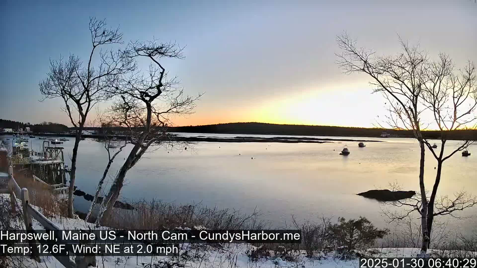 A calm, snow-dusted waterfront scene at sunrise shows several boats moored in a quiet harbor, with bare trees in the foreground and a soft, colorful sky.