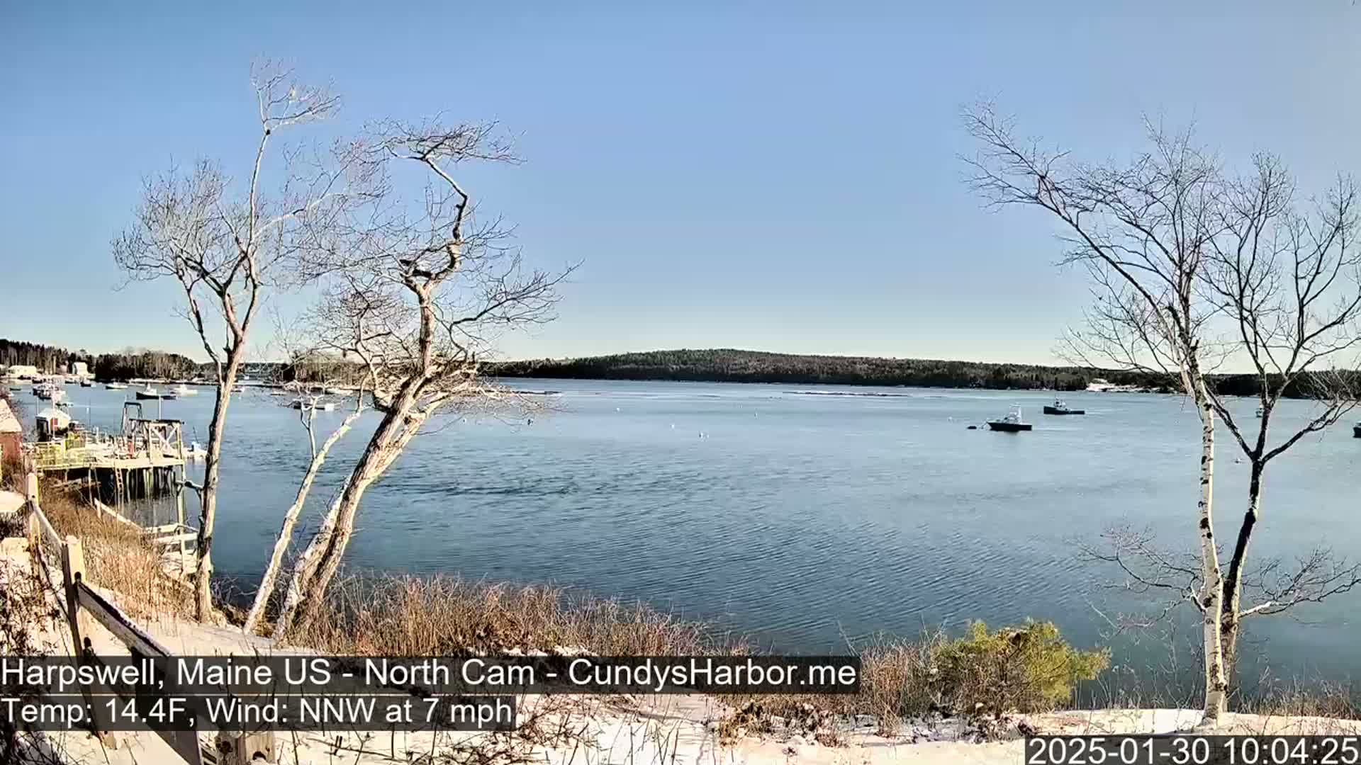 A calm body of water with a few boats, viewed from a snow-dusted shoreline with bare trees under a clear blue sky.