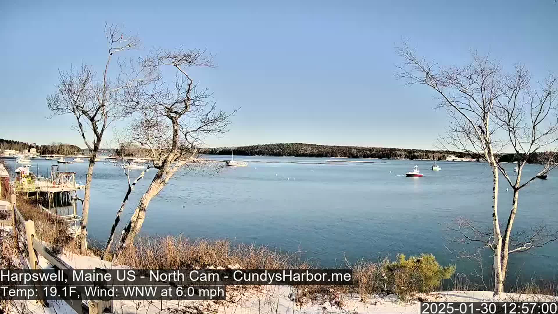 A calm body of water with several boats, viewed from a snow-dusted shoreline with bare trees under a clear blue sky.