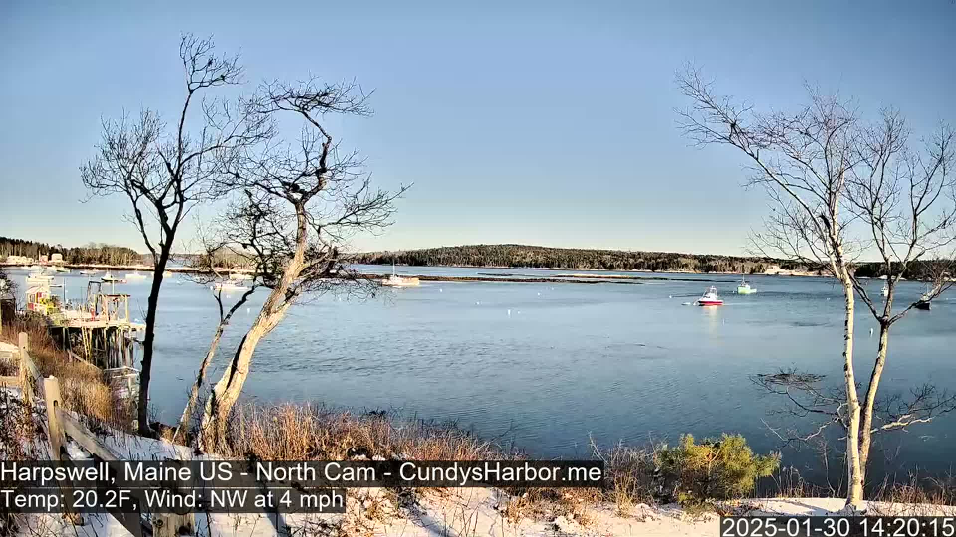 A calm body of water with several boats is seen from a snow-dusted shoreline under a clear, bright sky.