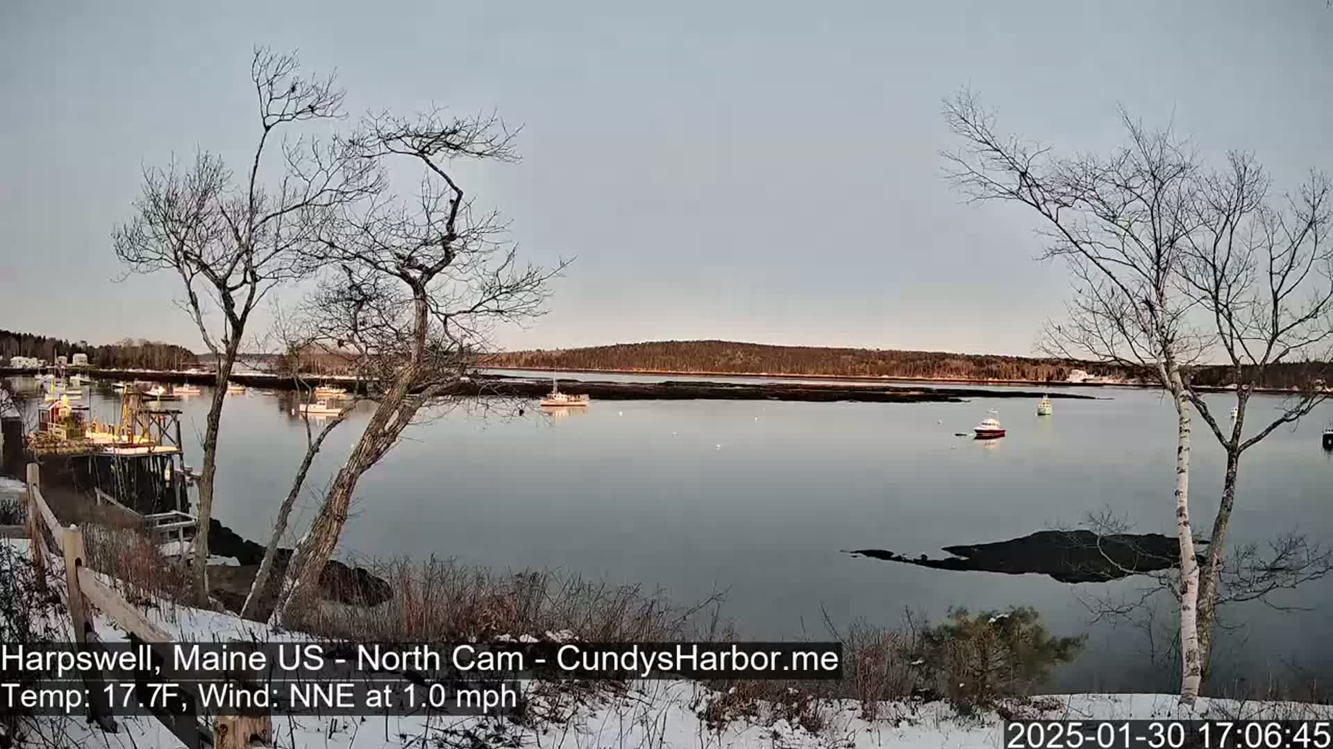 A calm body of water with several boats is seen from a snowy shoreline under a clear, light-grey sky.