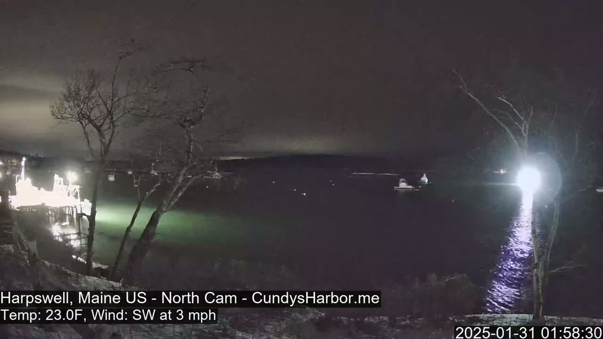 A nighttime view of a dark body of water with several boats, illuminated structures, and trees on the shore under a clear sky, with a southwest wind at 3 mph and a temperature of 23°F.