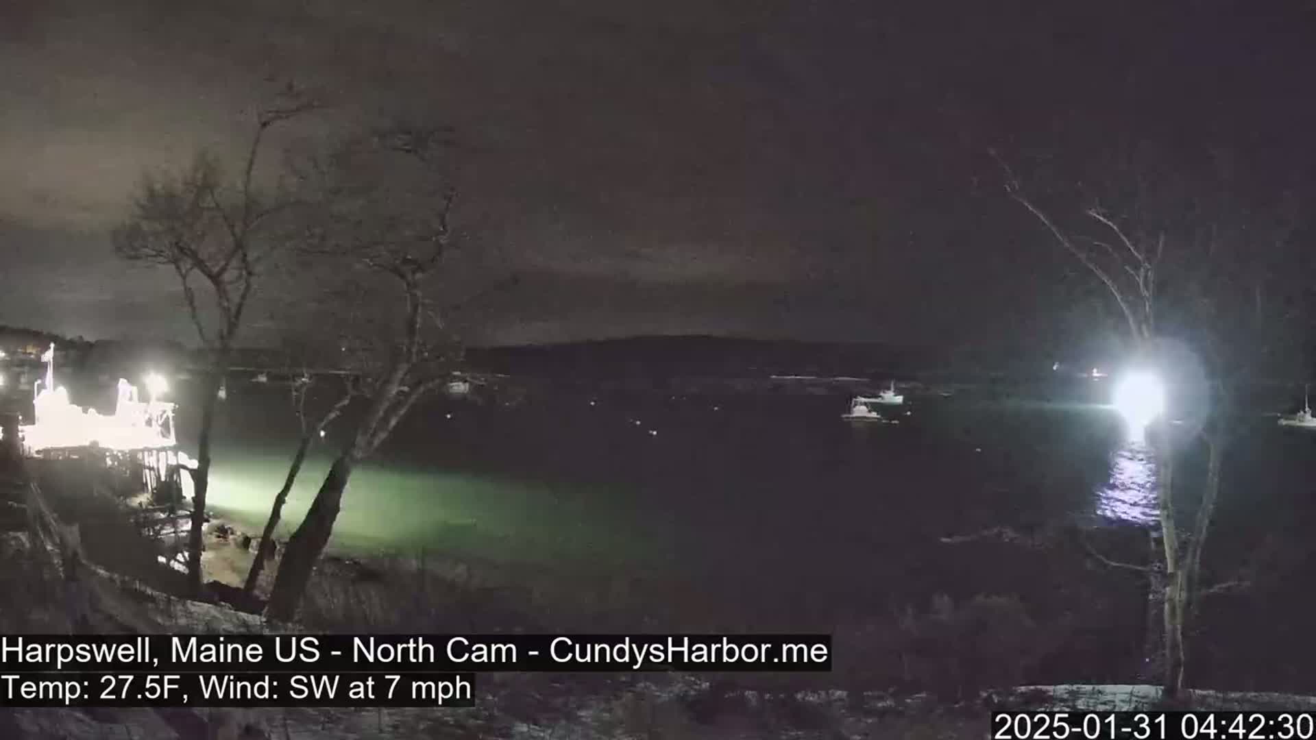 A nighttime view of a dark body of water with several boats, a brightly lit dock, and bare trees under a dark, clear sky, with a southwest wind at 7 mph and a temperature of 27.5°F.
