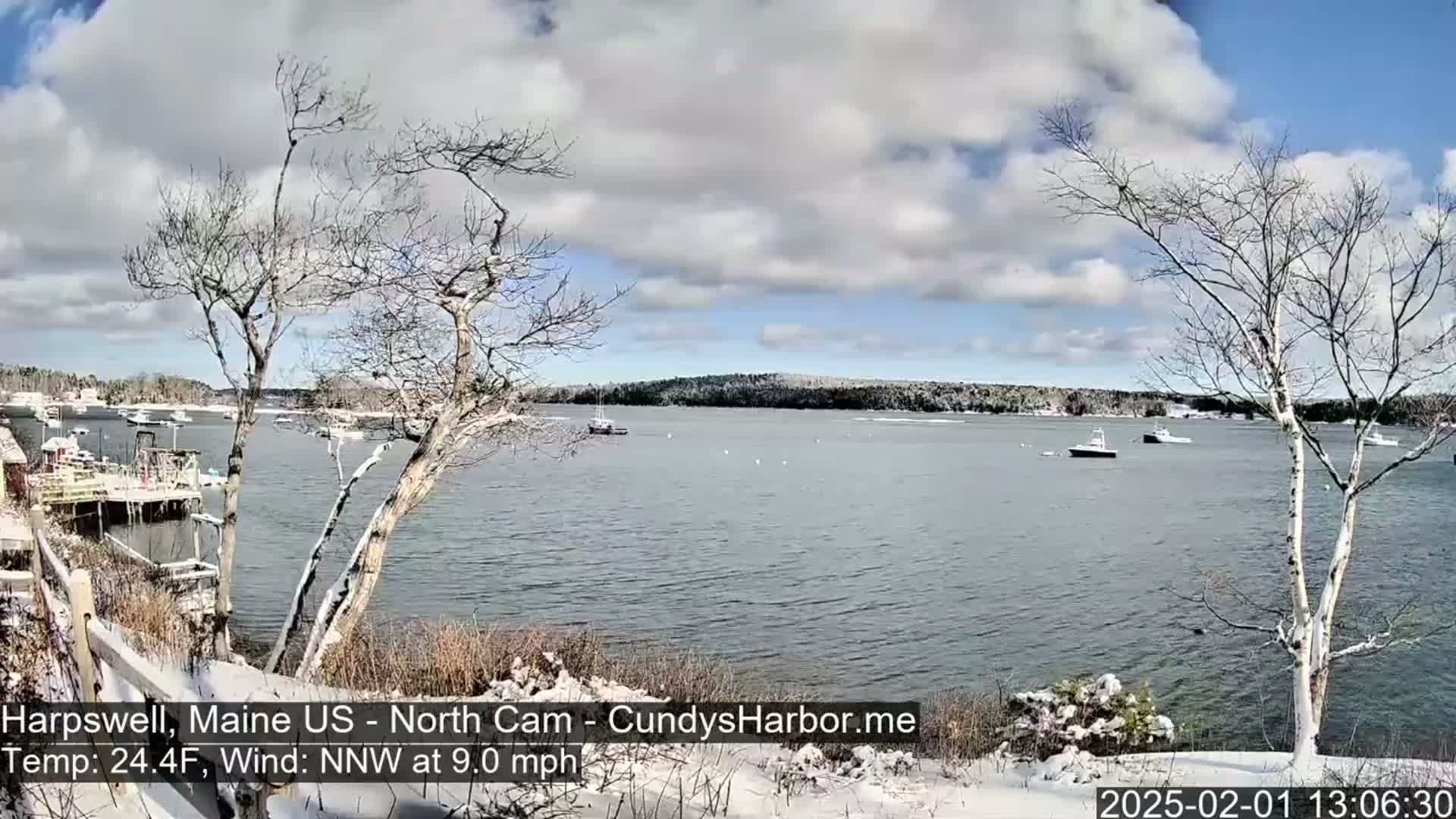 A snow-dusted shoreline overlooks a calm bay with several boats, under a partly cloudy sky.