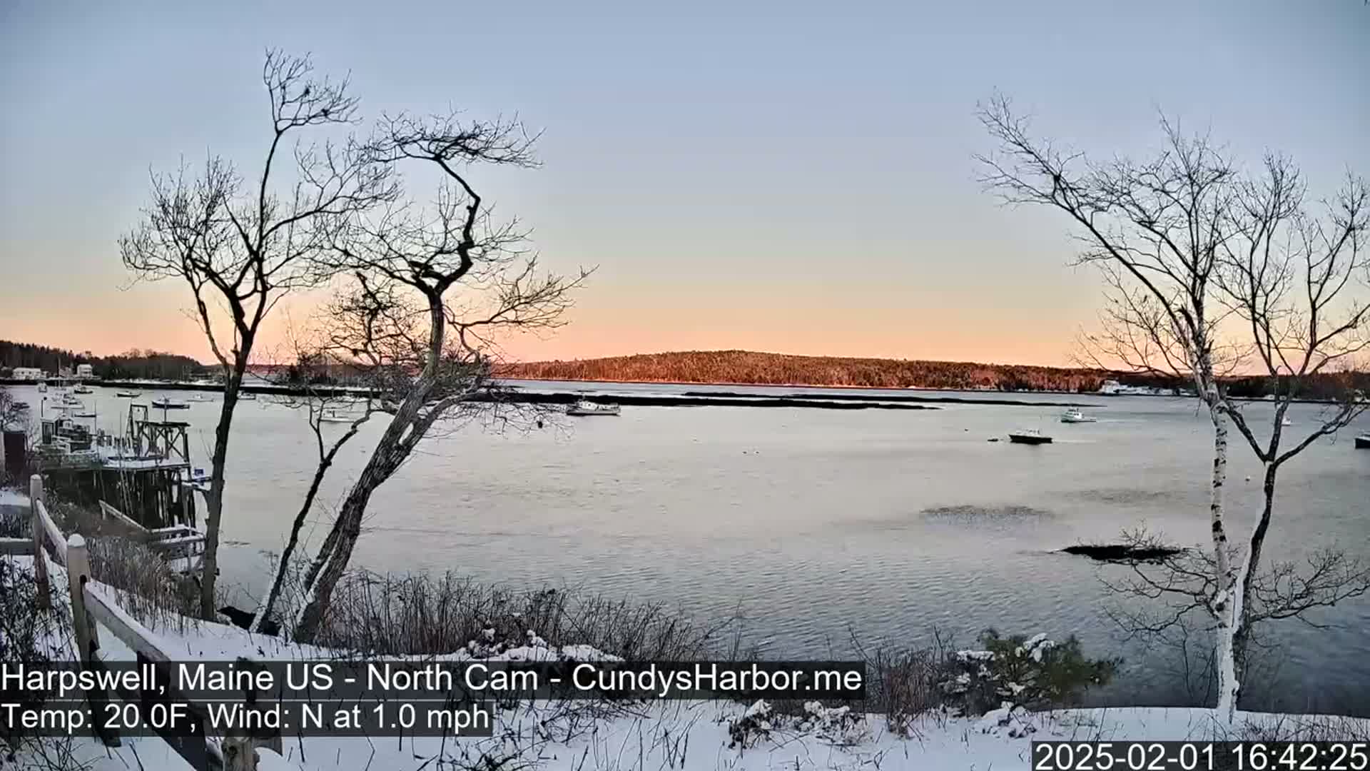 A snow-covered shoreline overlooks a calm bay with several boats, under a twilight sky.