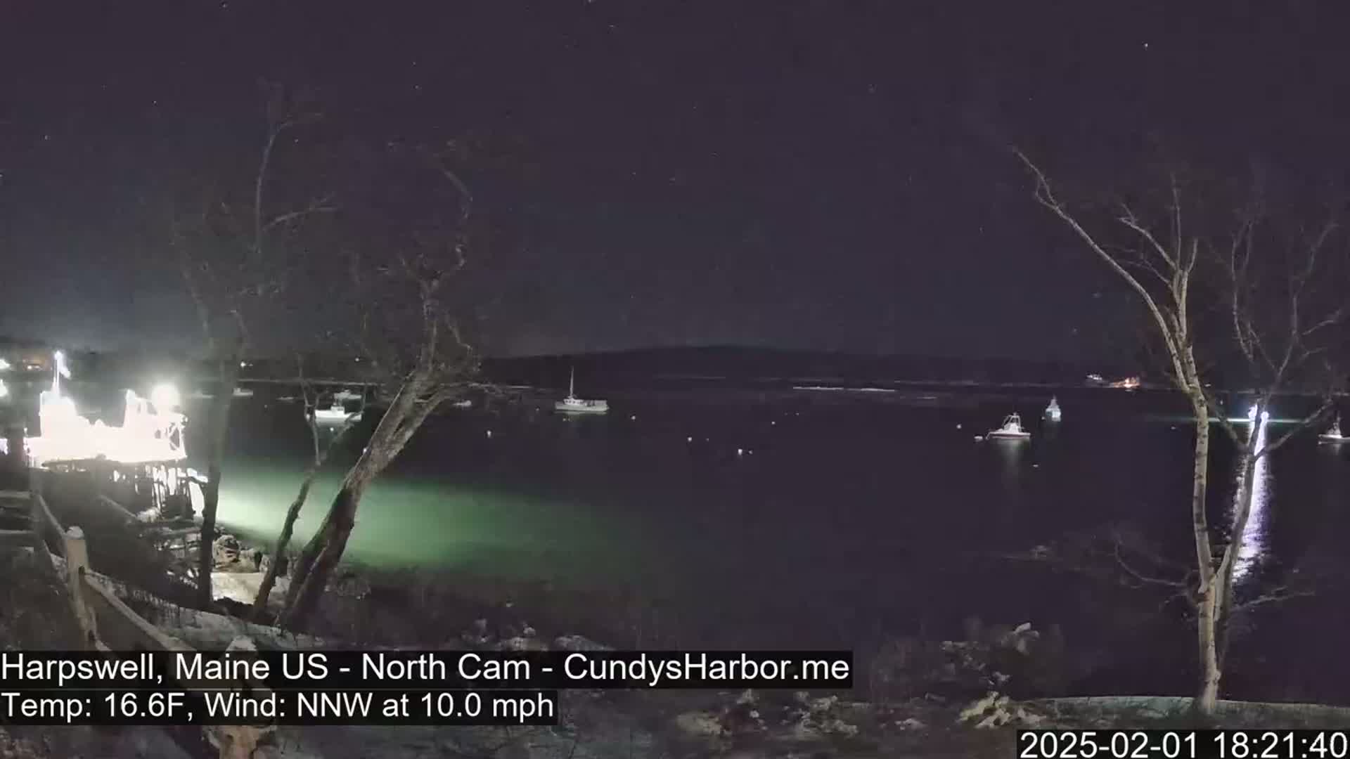 A nighttime view of a harbor with several boats, illuminated by lights from a nearby structure, under a clear, starry sky with a 10 mph northwest wind and a temperature of 16.6°F.