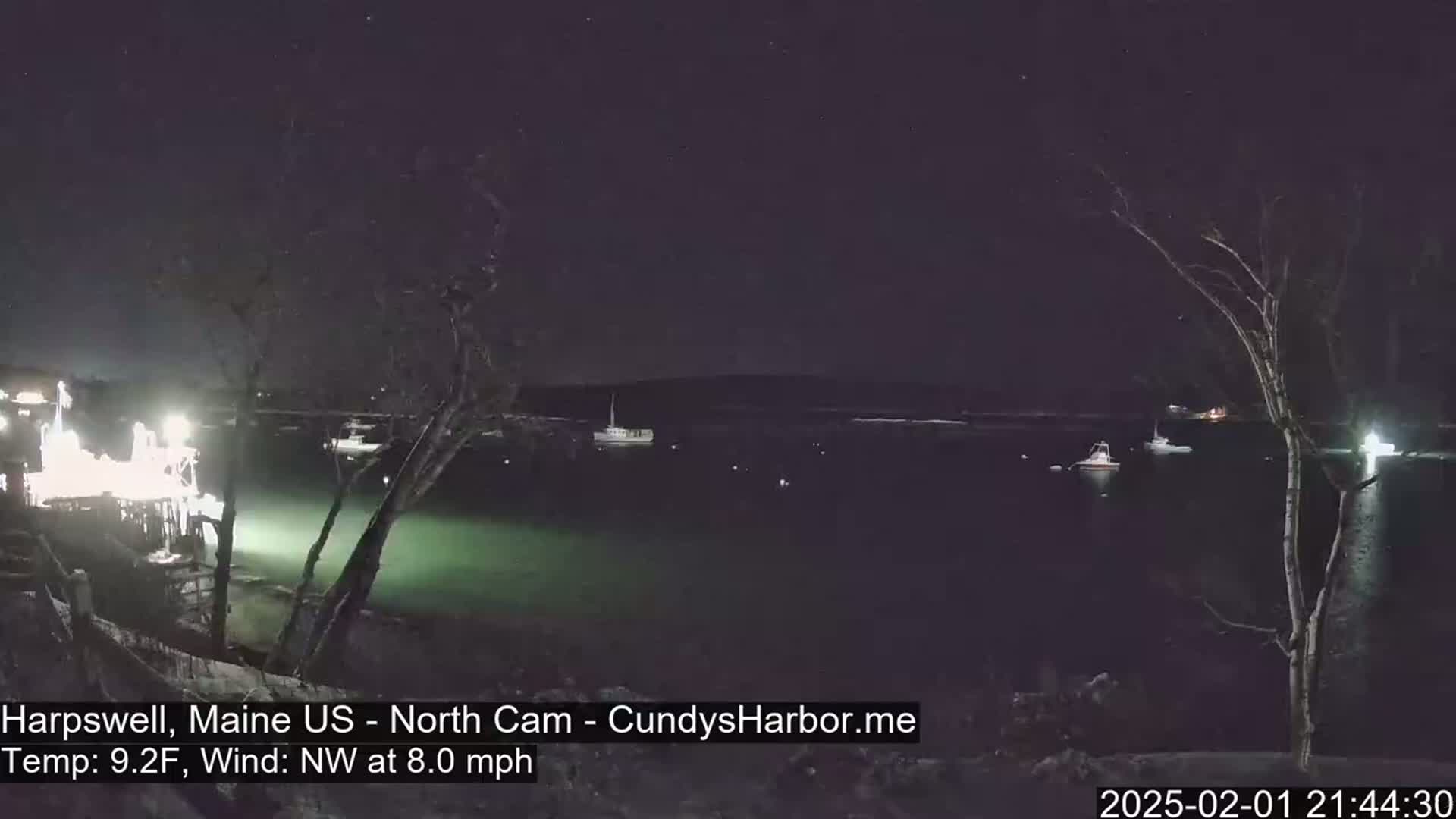 A nighttime view of a dark, calm body of water with a few boats, a shoreline with trees and lights, under a clear, starry sky.