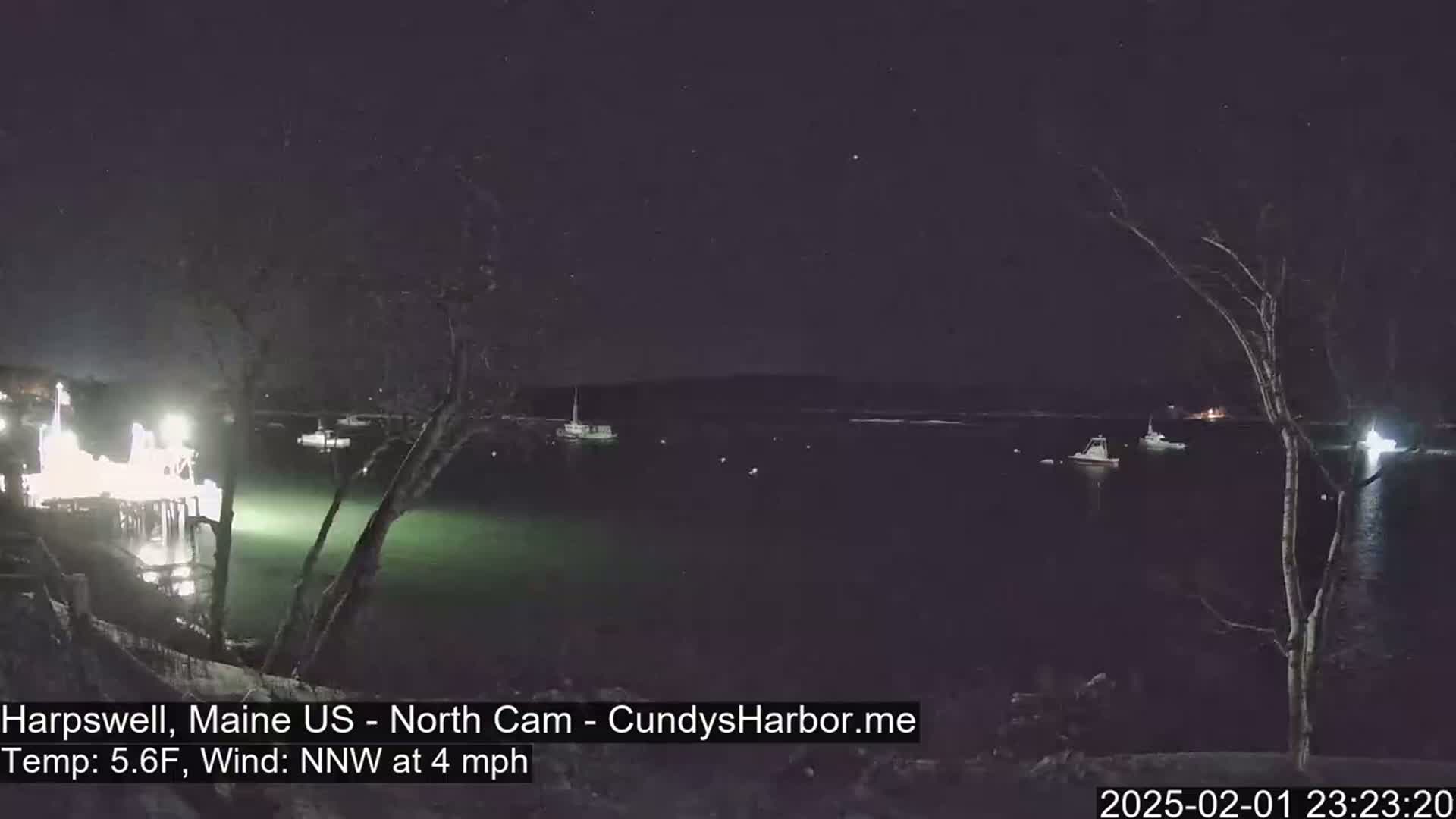 A nighttime view of a harbor with several boats, a dock with lights, and bare trees under a clear, starry sky, with a temperature of 5.6°F and a north-northwest wind at 4 mph.