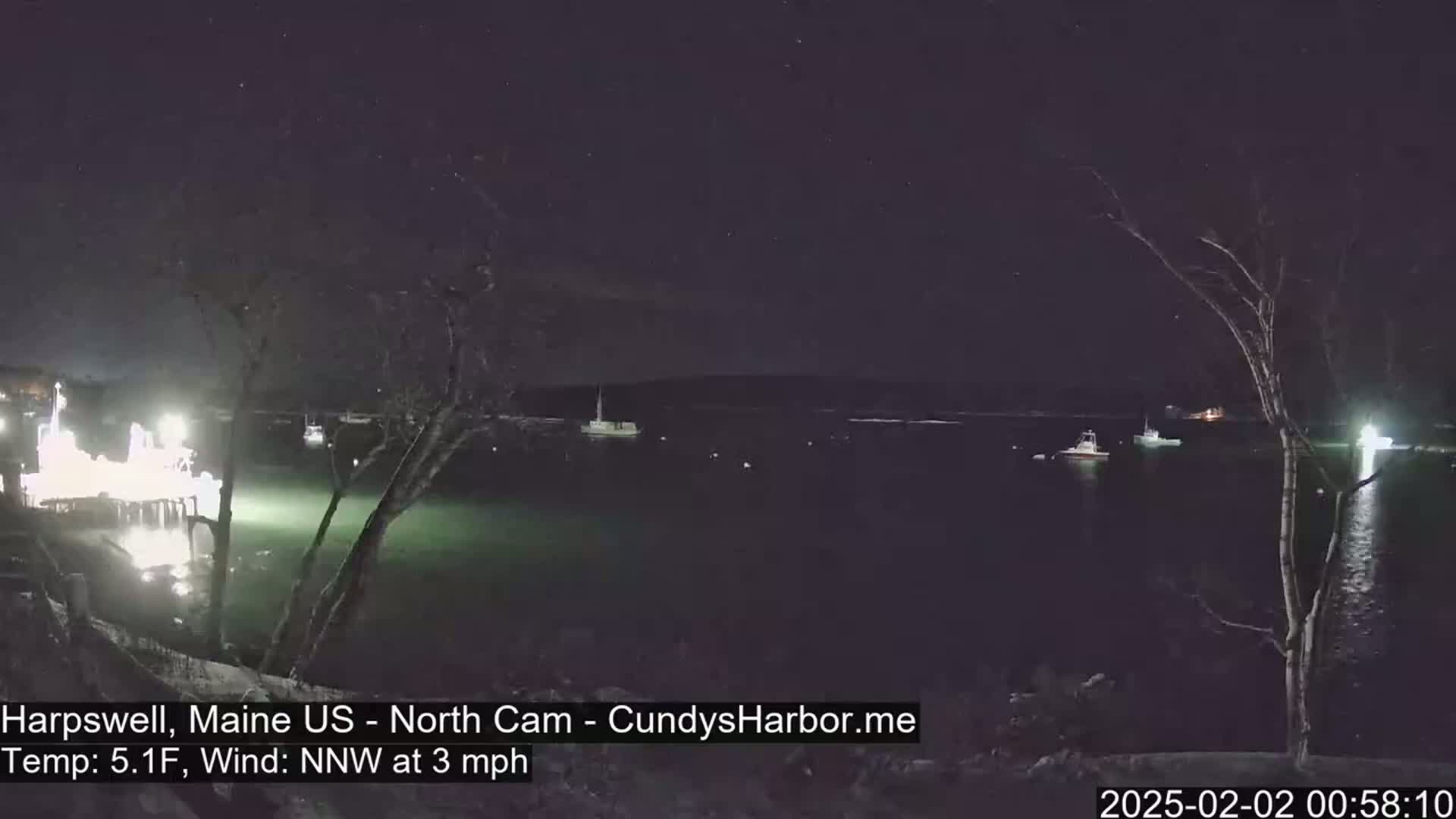 A nighttime view of a harbor with several boats, lit docks, and bare trees under a clear, starry sky; the temperature is 5.1°F and the wind is from the northwest at 3 mph.