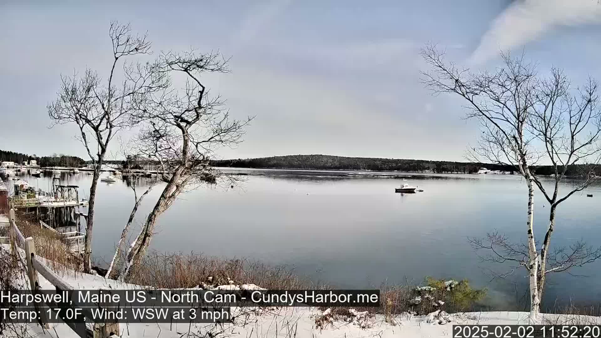 A snow-dusted shoreline overlooks a calm bay with a few boats, under a partly cloudy sky.