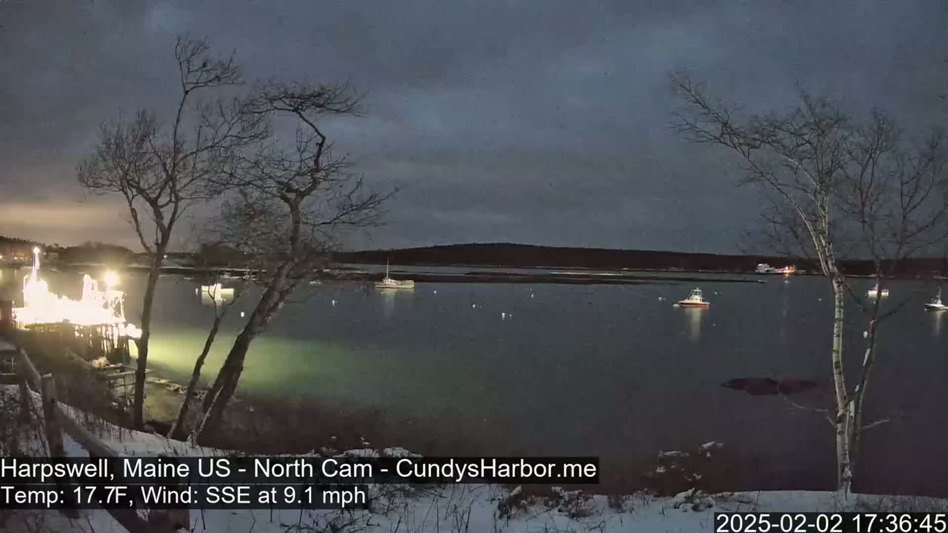 A nighttime view of a calm body of water with several boats, illuminated by lights on a nearby dock, under a dark, cloudy sky with a slight SSE wind.