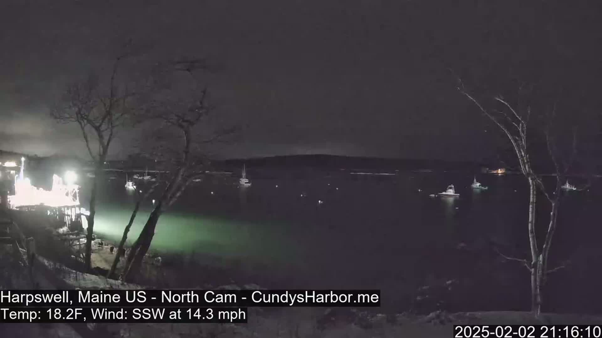 A nighttime view of a dark harbor with several boats, illuminated by lights on land, under a dark sky with a strong south-southwesterly wind.