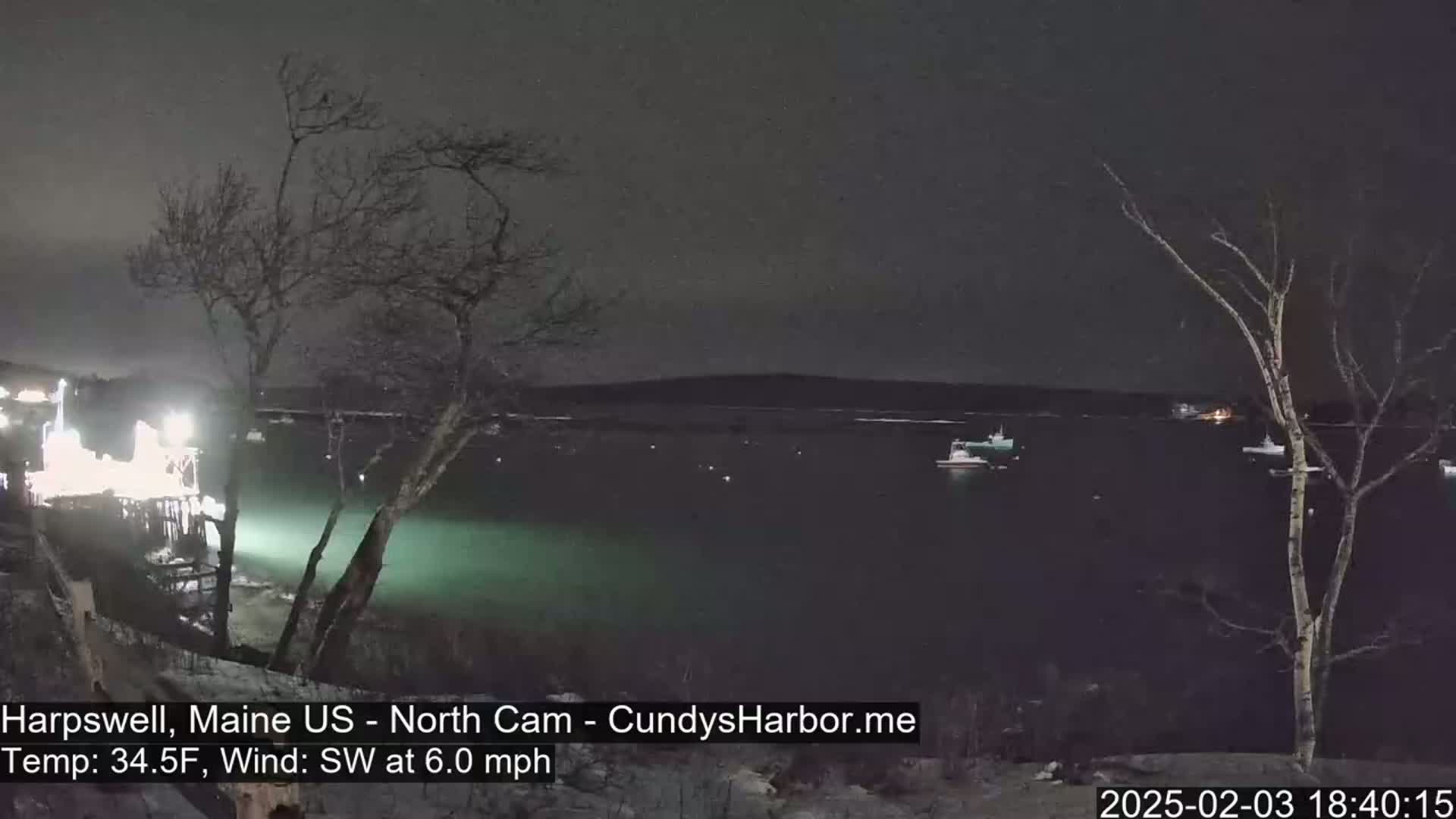 A nighttime view of a dark body of water with a few boats, under a mostly clear sky, with bare trees in the foreground and a slight southwest wind.