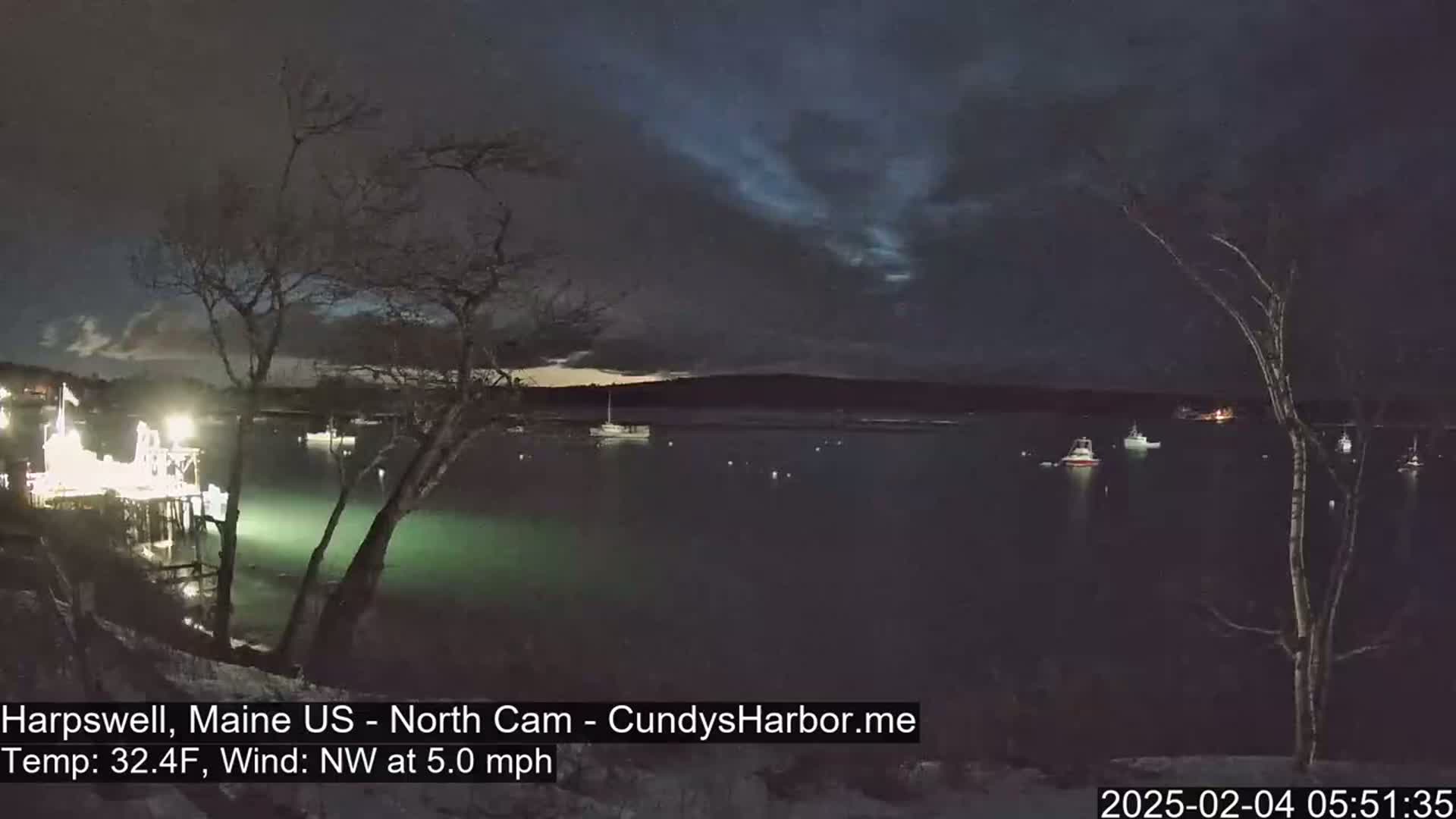 A nighttime view of a calm harbor with several boats at anchor under a dark, cloudy sky, with a temperature of 32.4°F and a northwest wind of 5.0 mph.