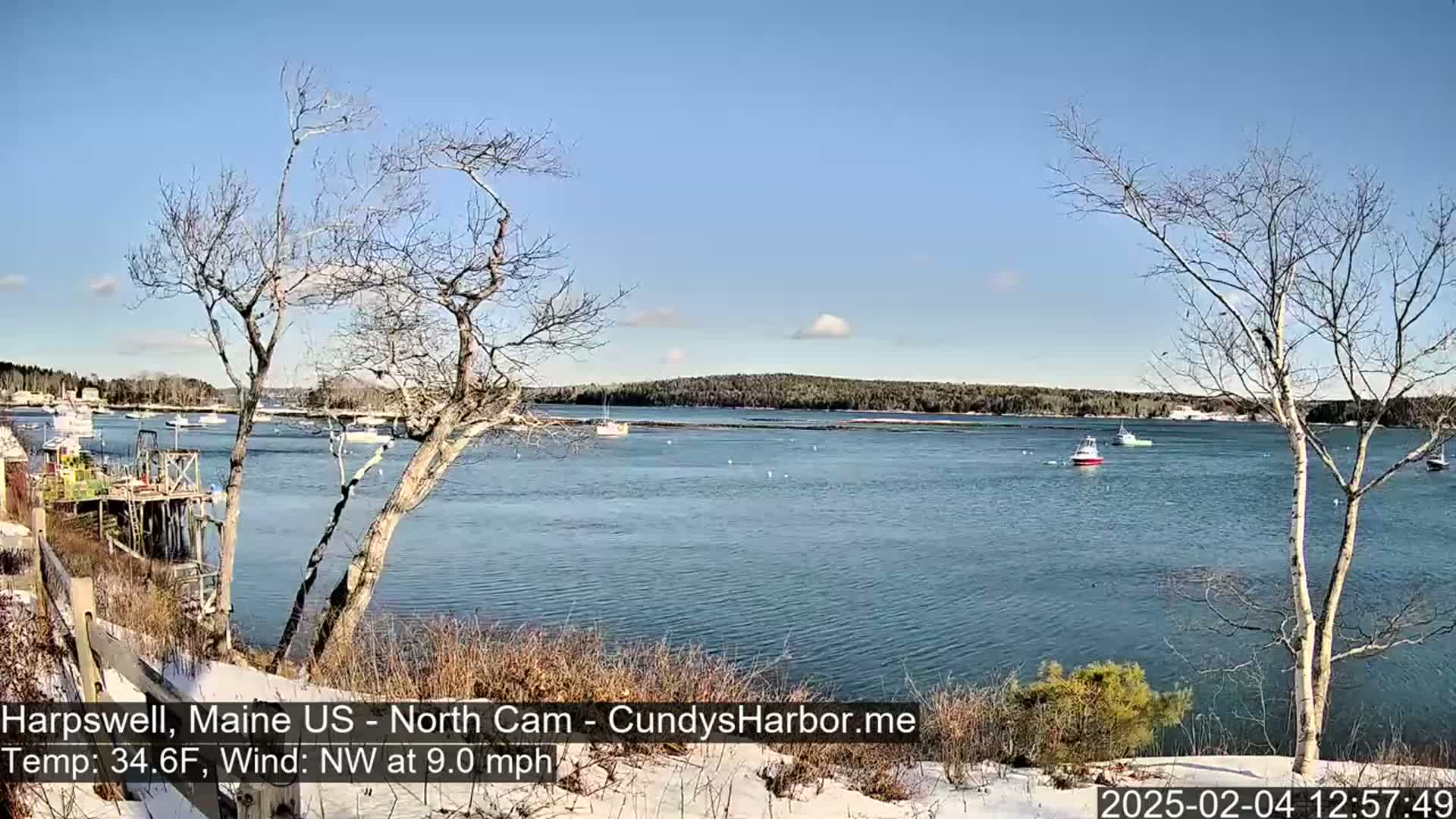 A tranquil bay with several boats is viewed from a snow-dusted shoreline on a sunny day with a northwest wind.