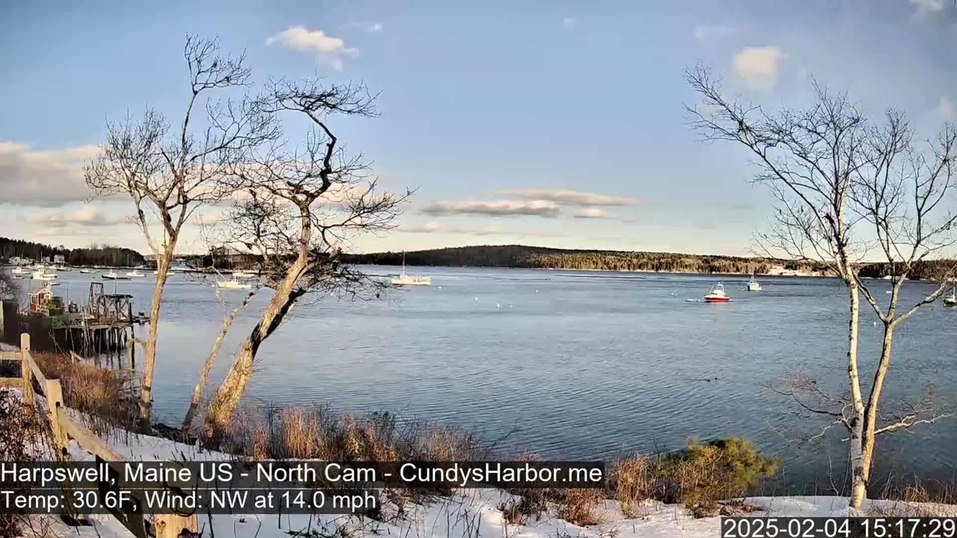 A partly cloudy, cool day with a northwest wind shows a calm harbor with several boats, viewed from a snow-dusted shoreline with bare trees.