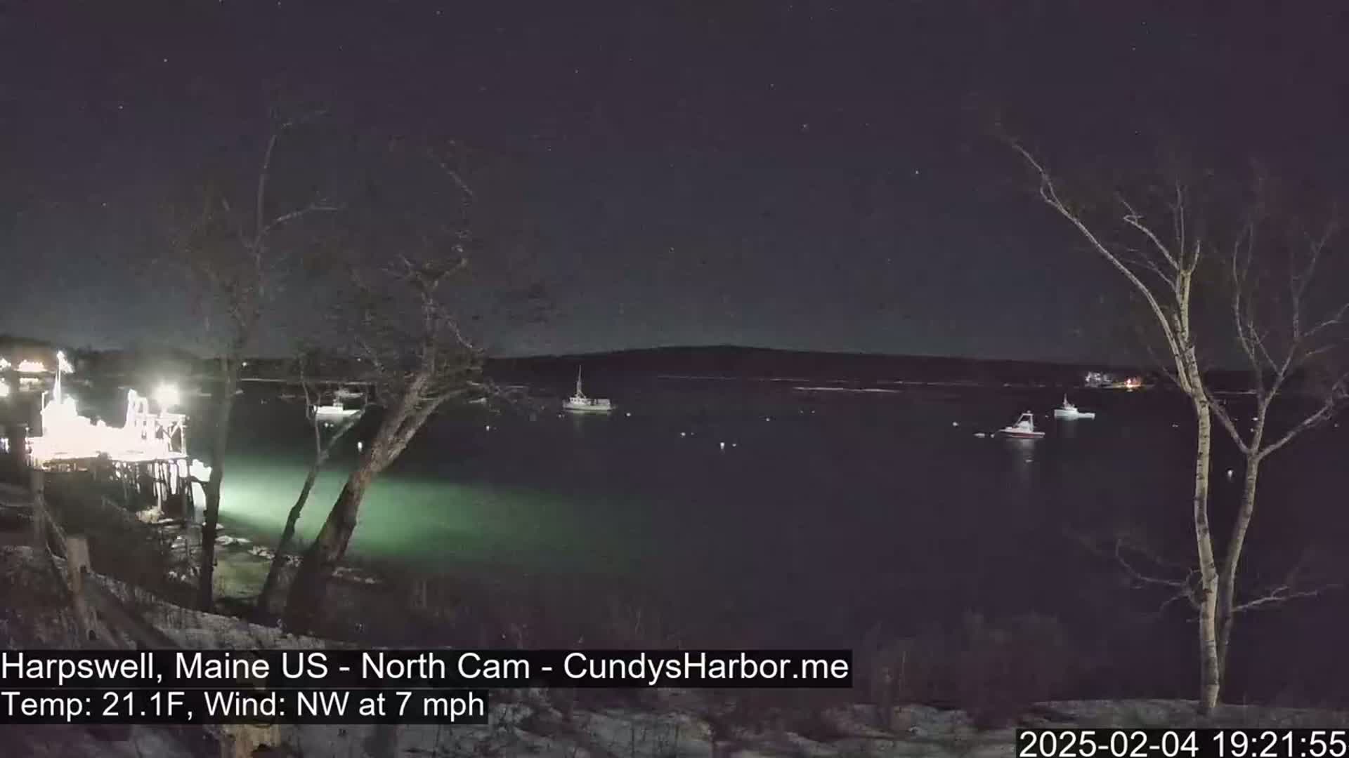 A nighttime view of a calm body of water with several boats, under a starry sky and a northwest wind at 7 mph.