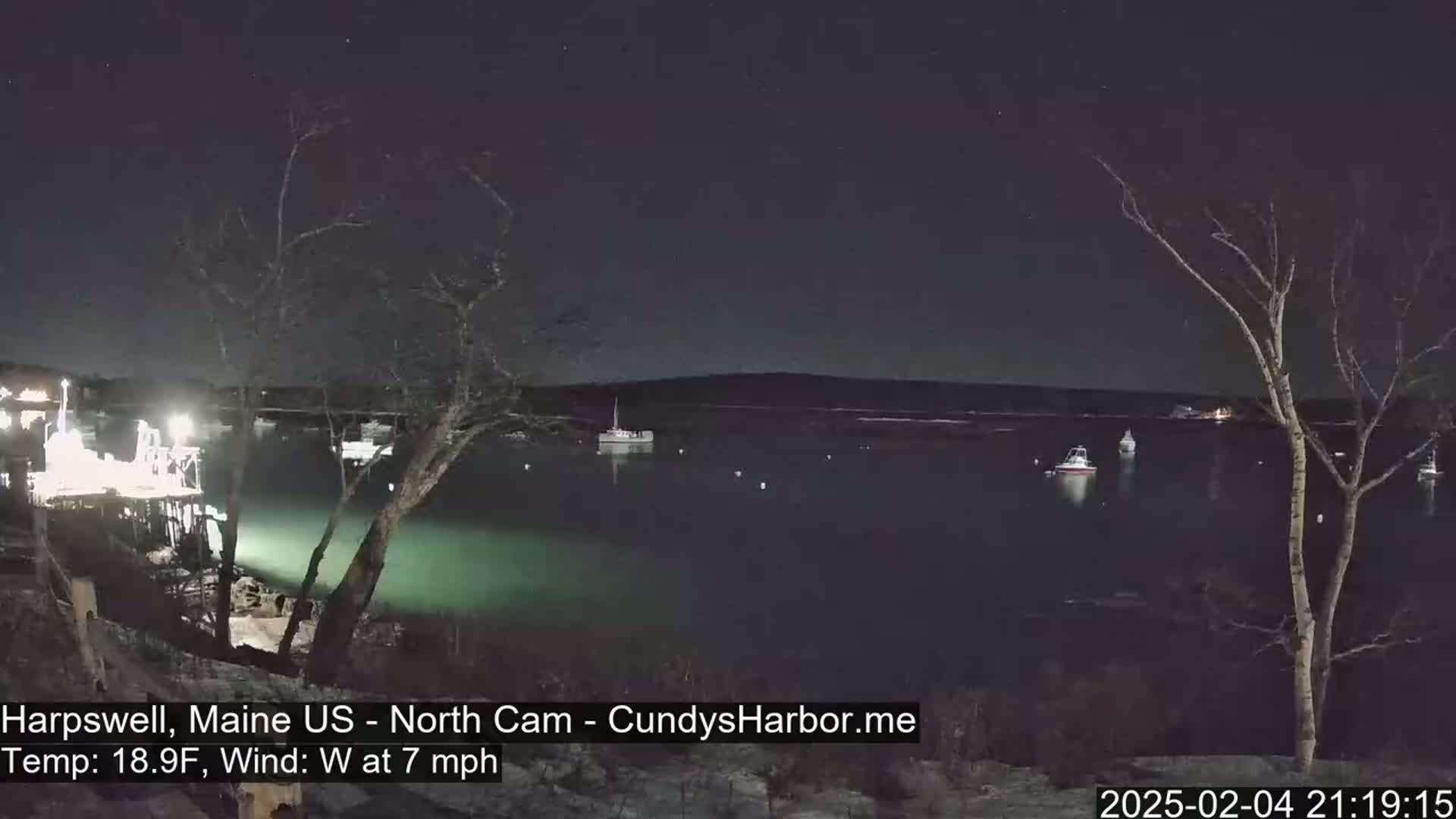 A nighttime view of a calm body of water with several boats moored, under a clear, starry sky, and a light westerly wind.