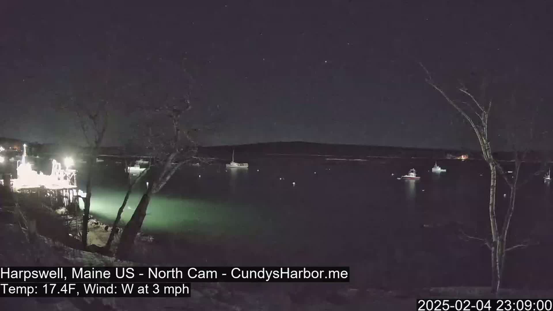 A nighttime view of a calm harbor with several boats, under a starry sky, and a temperature of 17.4 degrees Fahrenheit with a westerly wind of 3 mph.