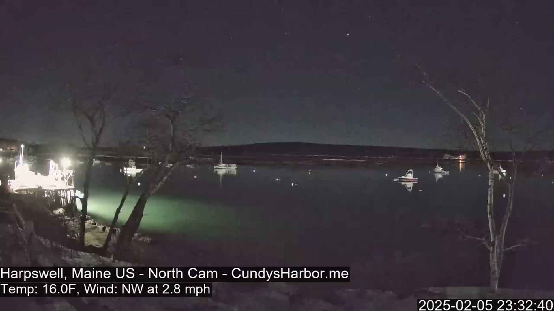 A nighttime view of a calm body of water with several boats moored, under a clear, starry sky, with a light northwest wind and a temperature of 16 degrees Fahrenheit.