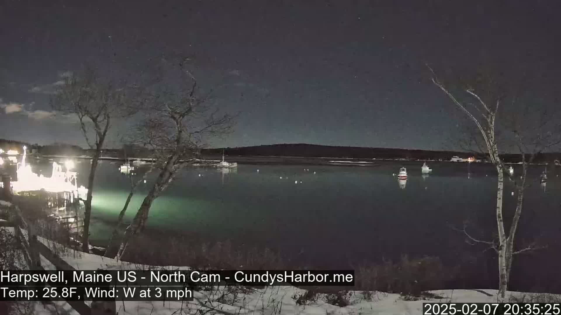 A nighttime view of a calm body of water with several boats, illuminated by lights from a nearby dock, under a clear, starry sky; there is snow on the ground.